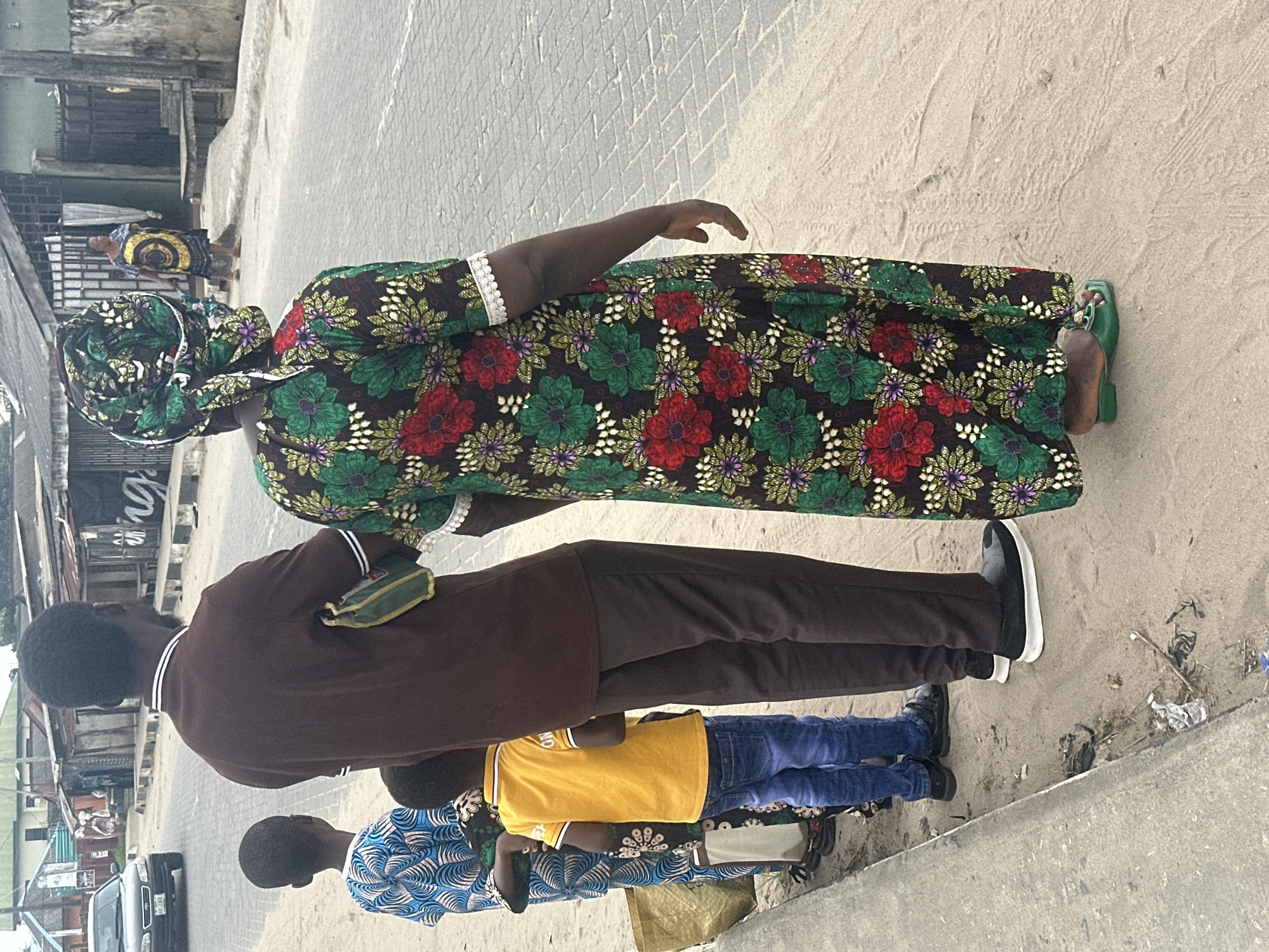In Badagry, Nigeria, a group of individuals stands near a dusty roadside with buildings in the background. A man wearing a brown shirt and dark pants stands in the center, facing away from the camera. To his left, a child in a blue and white patterned garment and another child in a yellow shirt and blue jeans are also facing away. To the right of the man, a woman in a vibrant green, red, and floral patterned dress with a matching headscarf is visible from the back. Another woman in a yellow and black patterned dress is further down the road. A black car is parked on the left side of the road. The environment appears to be an urban or semi-urban setting, with paved roads and a sandy shoulder.
