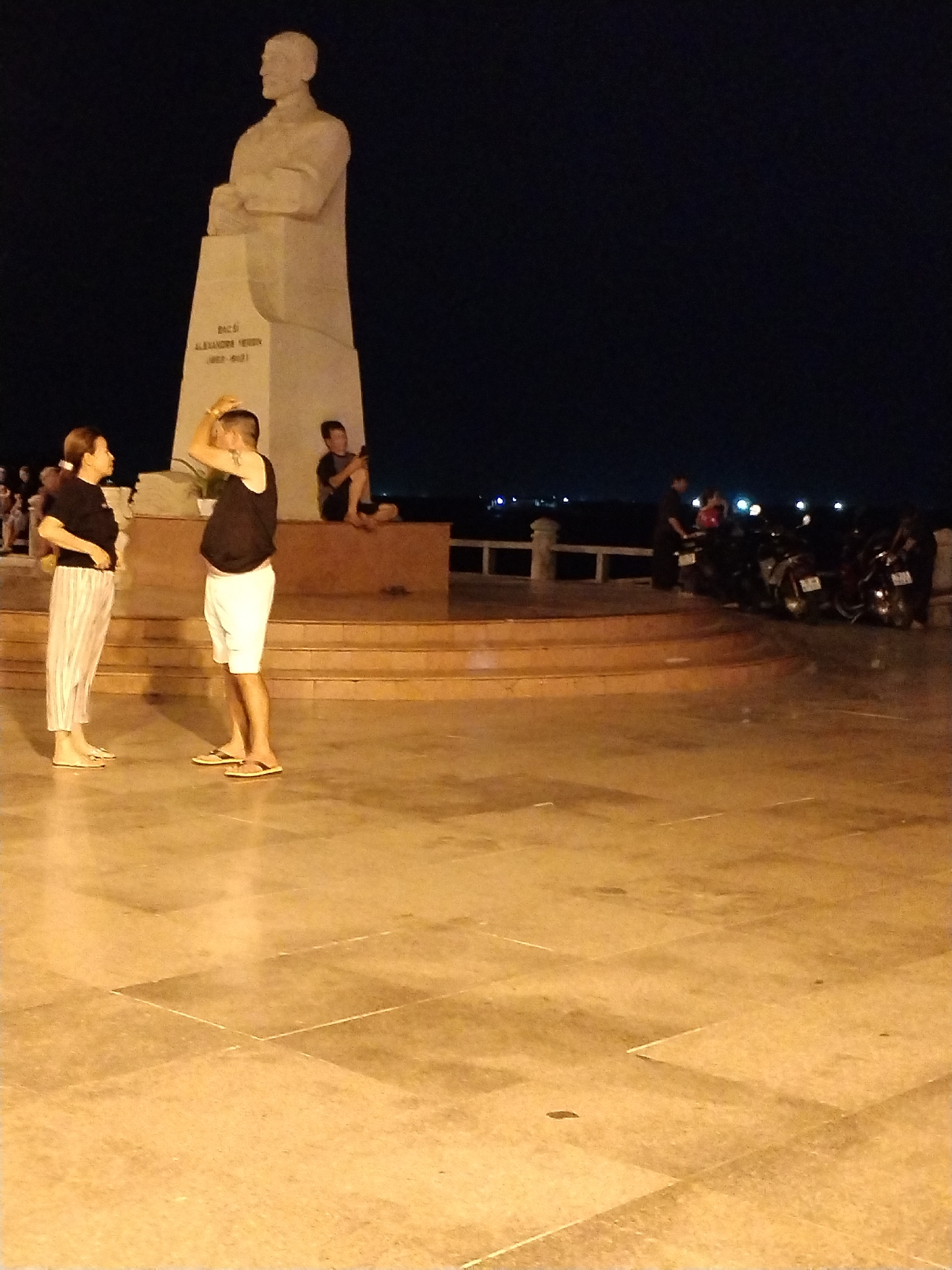 The image depicts a nighttime scene at what appears to be a public plaza in Nha Trang Ward, Vietnam, featuring a large statue of a historical figure on a tiered platform. The statue is inscribed with the name "BAC ALEXANDRU HERCULAN (1855-1941)". To the left of the statue, a woman wearing a dark top and striped pants stands facing a man in a dark tank top and white shorts who is raising his hands to his hair. A person is sitting on the edge of the platform behind them. Motorbikes and a few people are visible in the background near a seawall. The ground is paved with large tiles. The sky is dark.