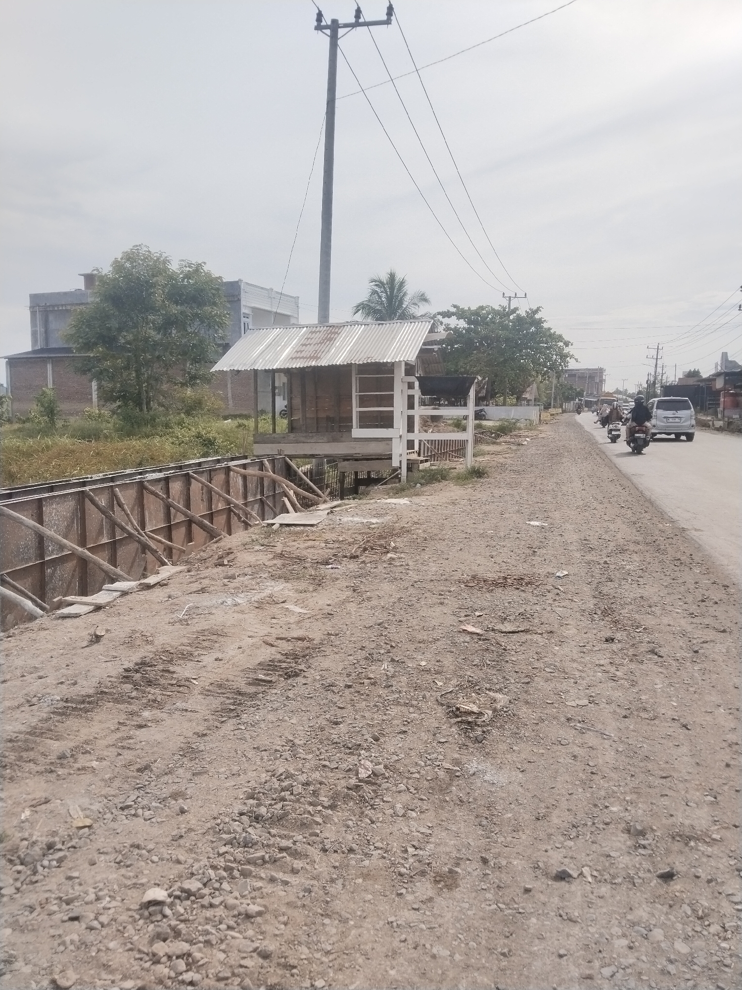 This daytime image captures a semi-urban or rural roadside scene in Barokulamgajah, Indonesia, under an overcast or hazy sky. In the foreground, a wide, unpaved dirt and gravel path stretches from the bottom left to the mid-right. To the left of this path, a long, deep ditch or canal is visible, lined with what appears to be wooden formwork and metal panels, supported by numerous wooden struts, suggesting ongoing construction or infrastructure work.
Adjacent to the ditch, a small, open-sided structure resembling a shack or stall stands, featuring a corrugated metal roof and white wooden framing. Further along, to the right of the dirt path, a paved road extends into the distance. Traffic is present on this road, including several motorbikes with riders (some wearing helmets) and a white minivan.
In the midground and background, a mix of buildings with simple architecture and various trees with green foliage line the road. Tall utility poles with multiple overhead power lines are prominent features against the light sky, indicating an established electrical infrastructure. No legible text is discernible on any objects, signs, or vehicles in the image. The scene depicts a functional but somewhat rustic environment with everyday activity.