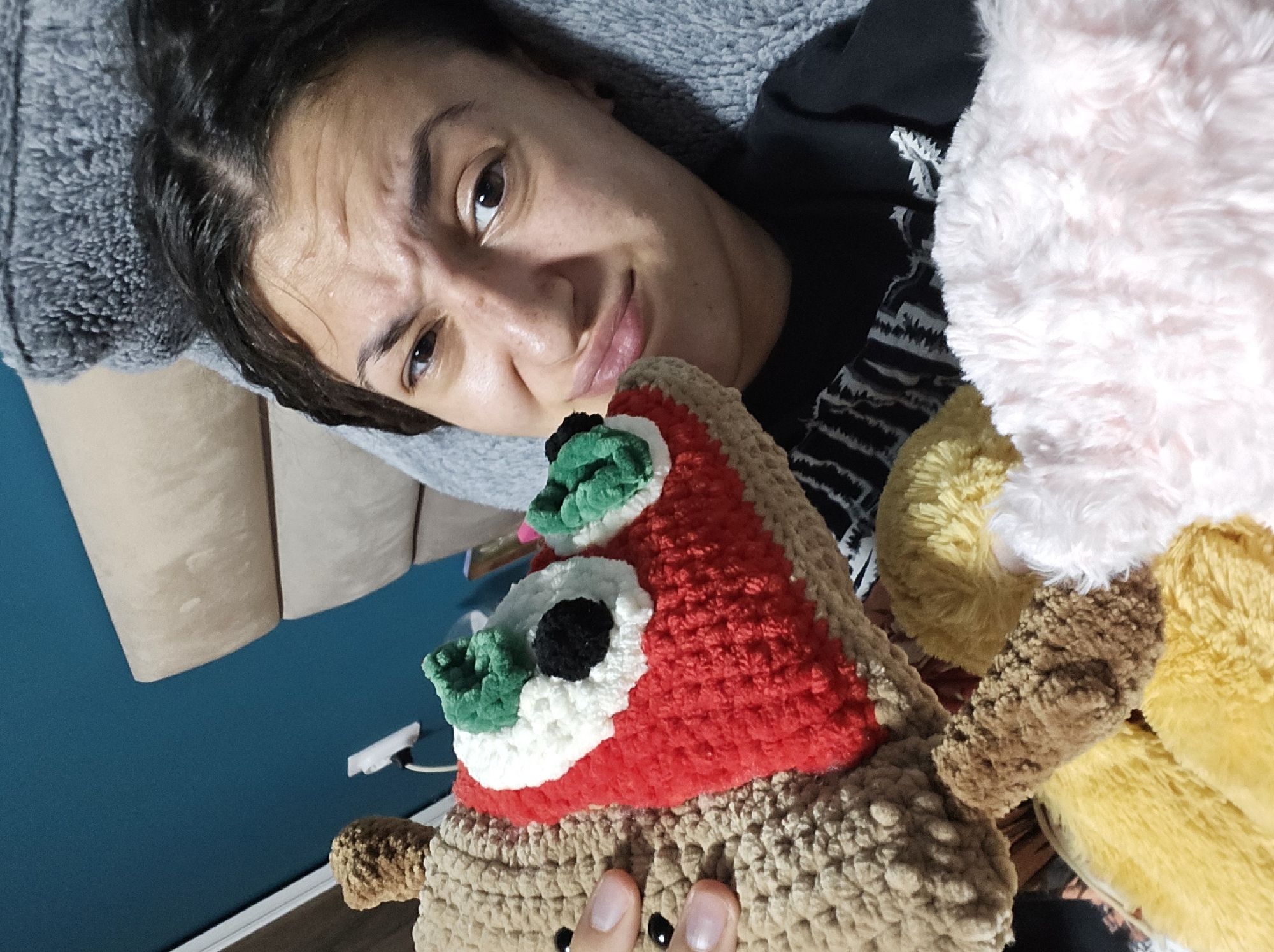 A woman is lying on a bed with gray bedding, holding a crocheted pizza slice. The pizza slice has a brown crust, red sauce, white cheese, and green and black pepperonis. To the right of the pizza slice are several other stuffed animals of various colors. The woman has dark hair and is looking at the camera with a slightly quizzical expression. The wall in the background is blue, and there are some beige objects on the left side of the frame. The location is not discernible from the image.