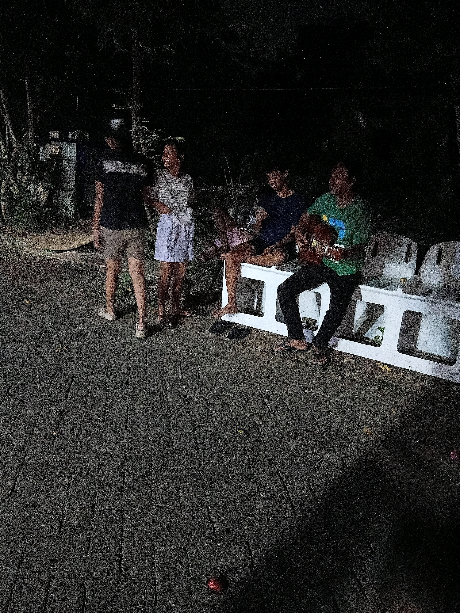 Four people are gathered outdoors at night, possibly in Cikal, Indonesia, near a paved walkway. Two individuals stand while two are seated. One standing person on the left wears a dark t-shirt and light-colored shorts. Beside them, another person stands wearing a striped t-shirt and light-colored shorts. To the right, a seated person in a dark t-shirt is partially visible. In front of the seated individuals, another person is seated and playing a guitar. They wear a green t-shirt and black pants. They are seated on a white plastic bench-like structure with other identical structures visible to their right. The background is dark with some foliage and what appears to be a building structure. The ground is paved with dark rectangular stones.