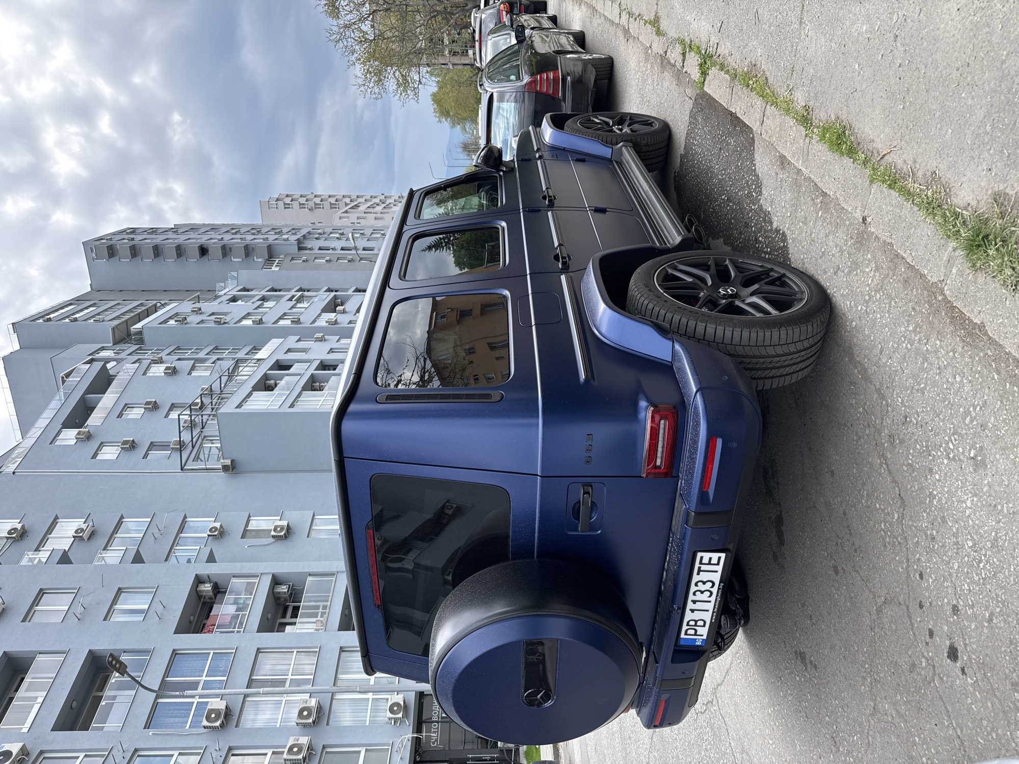 The image depicts a matte blue Mercedes-Benz G-Wagen (G63) parked on the side of an urban street during daylight hours, likely under an overcast sky. The luxury SUV is viewed from its rear and right side, with its rear wheels slightly turned. It features black multi-spoke wheels and a matching blue spare tire cover on the tailgate. Other cars are parked in a line further down the street behind it.
To the left of the parked vehicle, prominent light gray multi-story apartment buildings rise into the background. These buildings exhibit a modern, somewhat angular architectural style, characterized by numerous windows and visible external air conditioning units attached to their facades.
The ground consists of an asphalt street surface and a concrete curb, with sparse patches of grass and weeds growing along the edges. No people are visible in the scene.
Visible text includes the Bulgarian license plate "PB 1133 TE" with the country code "BG" and the website "www.bestinqrup.bg" printed below the number. The model designation "G63" is subtly embossed on the tailgate of the Mercedes. The scene suggests a typical street parking environment in a residential area of Plovdiv, Bulgaria.