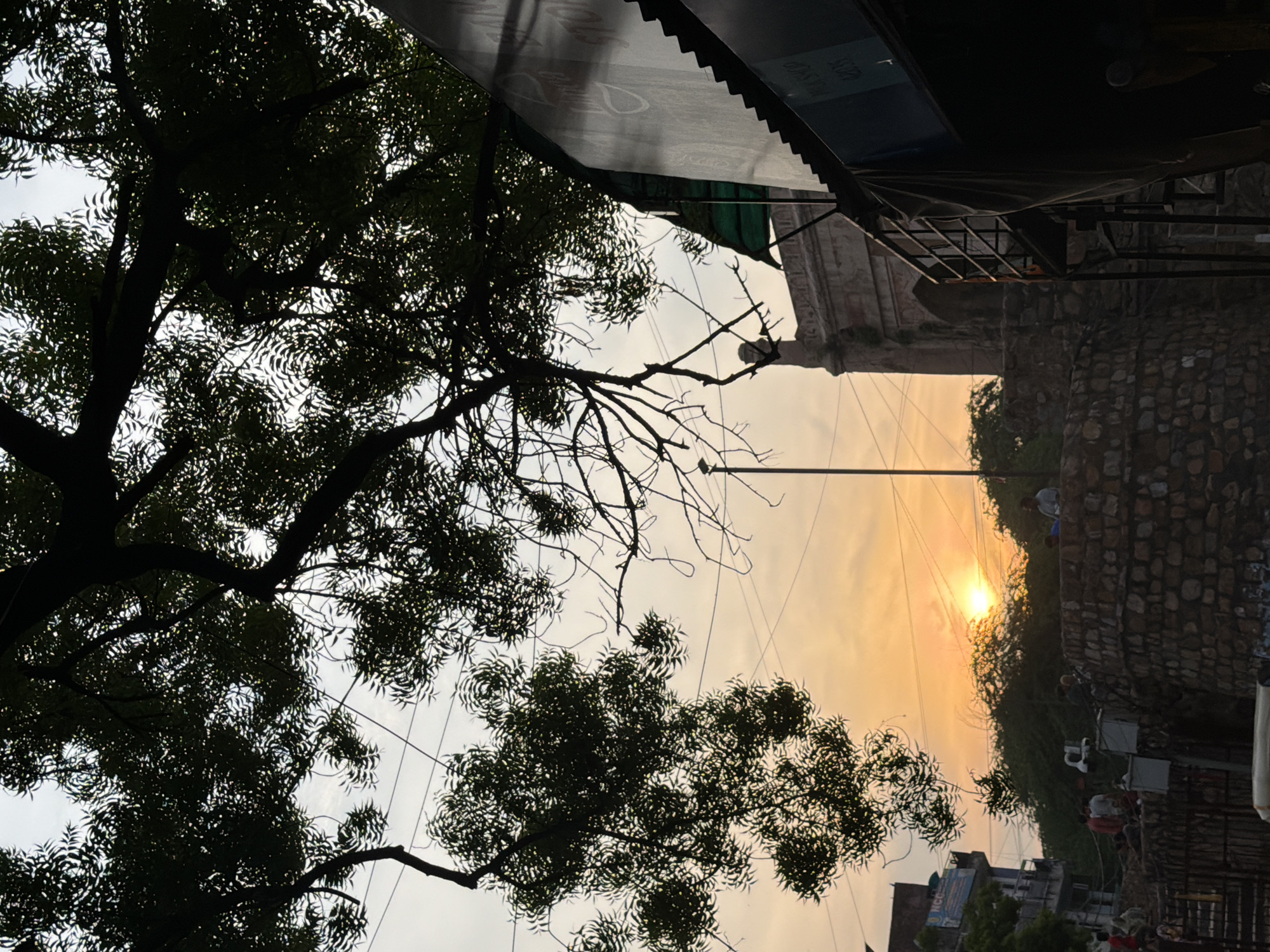 A sunset scene unfolds in Delhi, India, with the sun low on the horizon, casting an orange glow across the sky. Silhouetted trees dominate the foreground, their branches stark against the fading light. In the mid-ground, a stone wall curves around an area that appears to be an excavation site or a park. Several people are visible, some standing near the wall and others on what looks like stairs leading down. To the left, a multi-story building with balconies stands, and further back, a large gateway or structure with arched entrances is partially visible. On the right side of the image, a dark, sloped structure, possibly a shop awning, juts into the frame. Power lines crisscross the sky, a common sight in urban landscapes. The overall atmosphere is tranquil, tinged with the warm hues of dusk.