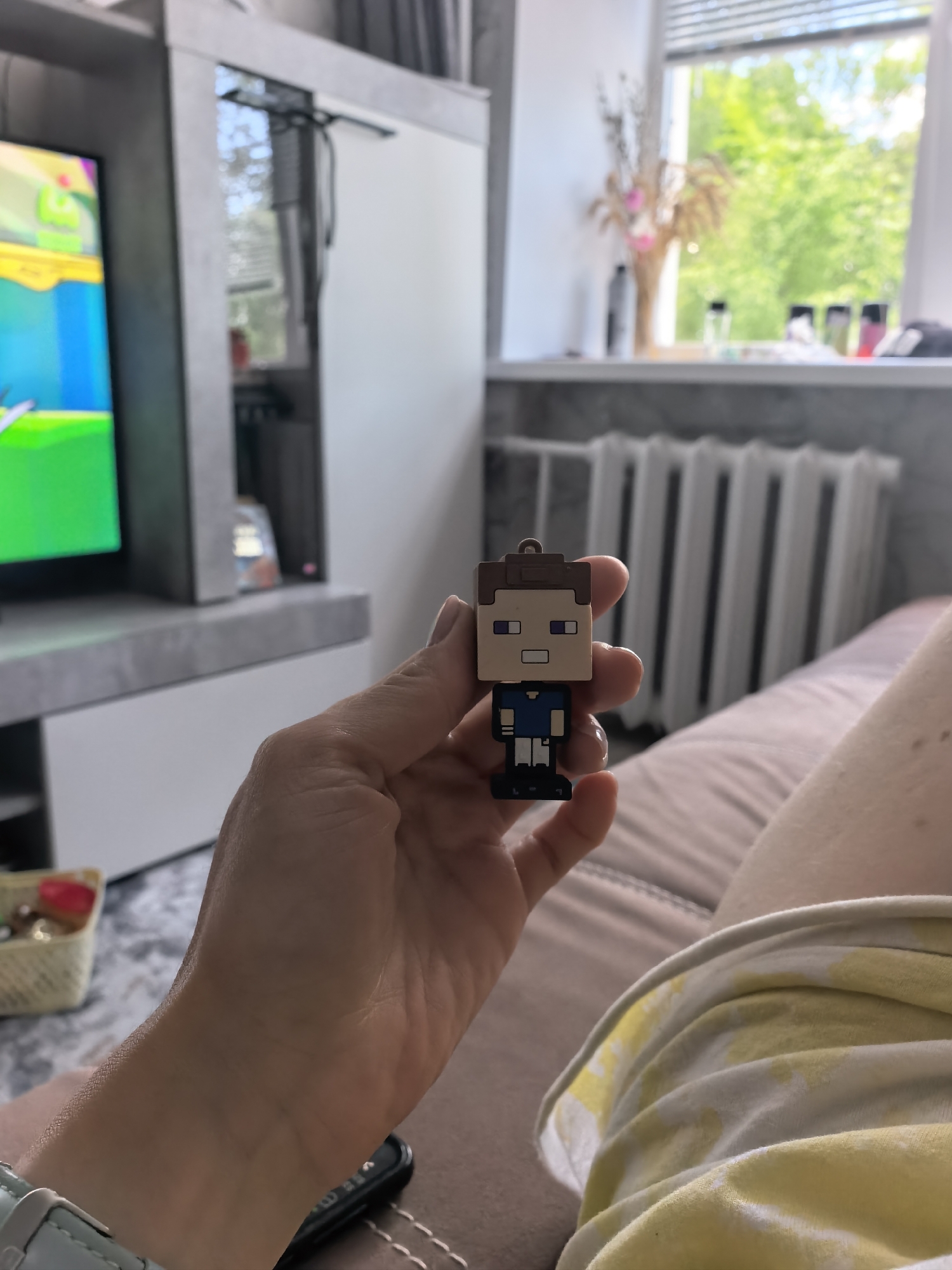 A person is holding a Minecraft Steve keychain in front of a living room setting. The background features a television set, a window with a view of greenery, and a radiator. The person is sitting on a couch, wearing a yellow and white shirt. A television cabinet is partially visible. The location is plausibly a residential interior. The location within the city of Sterlitamak, Russia, cannot be determined from the visual information provided.