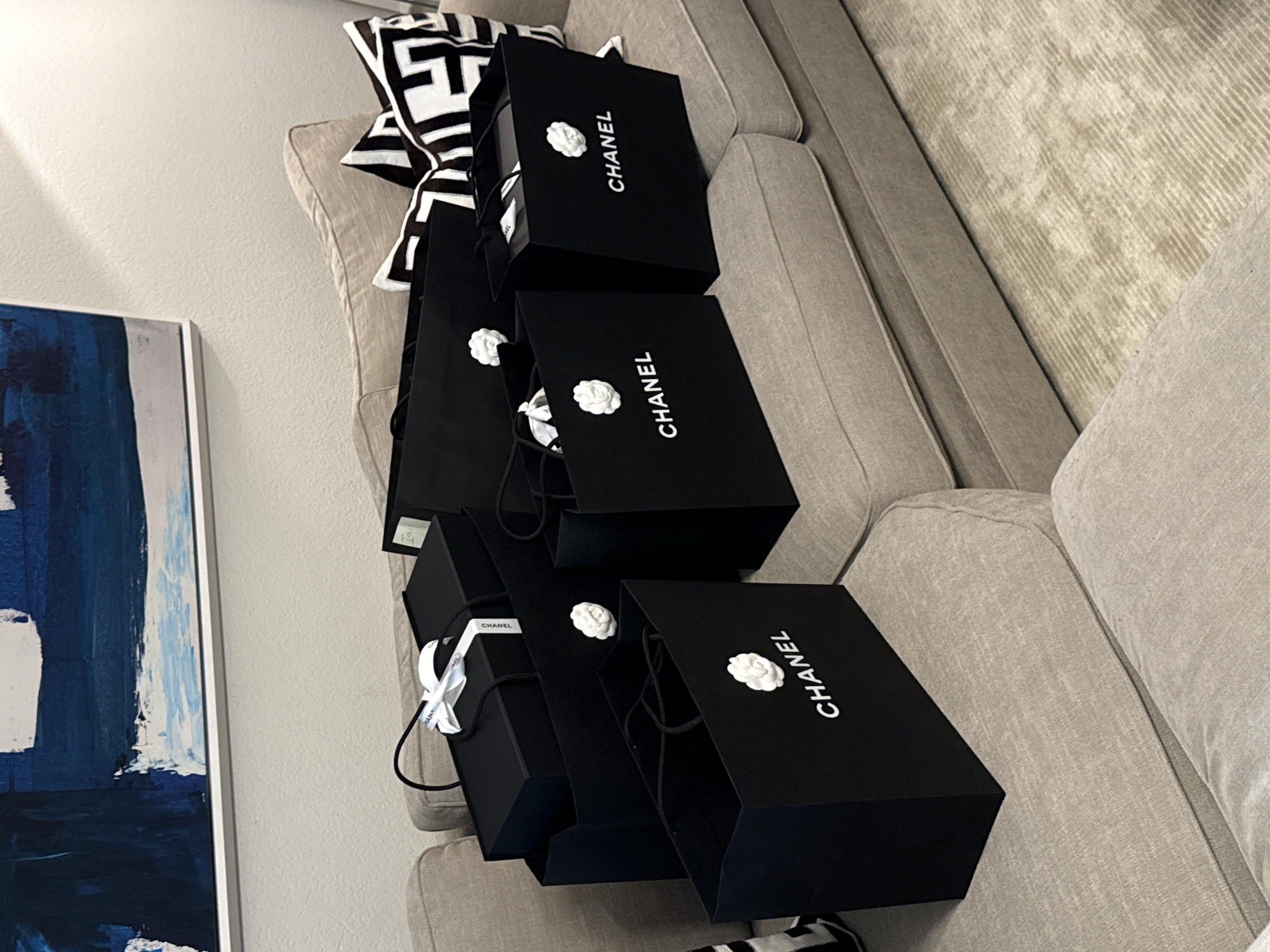 The image shows a stack of six black Chanel shopping bags on a light-colored upholstered sofa with a geometric patterned pillow behind them. The sofa appears to be located indoors, possibly in a residential setting, as evidenced by the framed artwork hanging on a wall. While the city of Chula Vista, United States of America, is specified, there is no visual information within the image to confirm or establish this location. The focus is solely on the stacked shopping bags and the immediate surrounding objects.