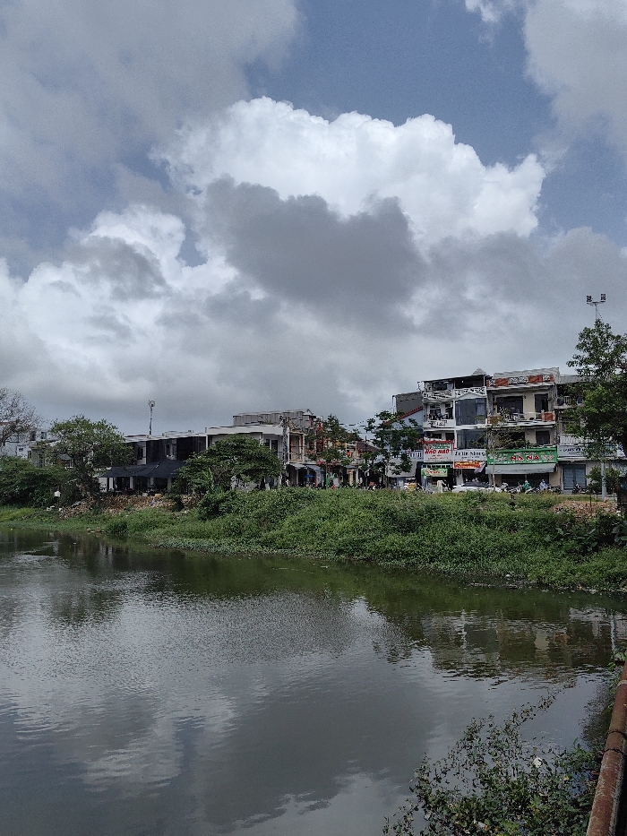 This image depicts a daytime scene in Phú Xuân Ward, Vietnam, viewed across a body of water towards an urbanized bank.
In the foreground, a dark green-brown body of water, likely a river or canal, occupies the lower half of the frame, showing ripples and reflections of the sky and buildings. Patches of green aquatic vegetation are visible on the water's surface and along the near bank, with a reddish-brown structure or railing partially visible in the bottom right corner.
The midground features the far bank, densely covered with varied green vegetation, including bushes and small trees. Behind this foliage, a row of multi-story buildings lines a street. These structures exhibit diverse architectural styles, with some featuring balconies and ground-floor storefronts displaying signs, such as "Mao" and "ANNA" partially legible. Several motorbikes are parked along the street in front of the shops, and a few indistinct human figures are present near the buildings. A light pole with multiple luminaires stands on the right side of the buildings, and a cellular antenna is visible on a rooftop in the mid-left.
The background consists of a partly cloudy sky, featuring large, scattered white and gray cumulus clouds against patches of light blue sky. The lighting suggests ambient daylight conditions.