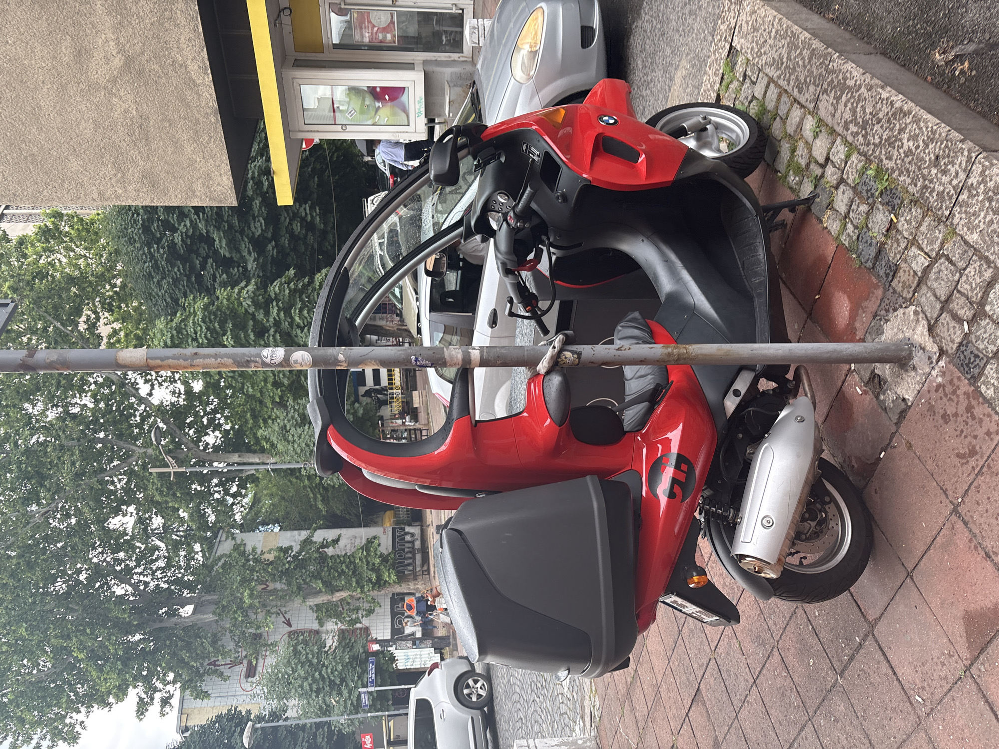 A red and black three-wheeled scooter is parked on a sidewalk next to a metal pole in Belgrade, Serbia. The scooter has a covered storage compartment on the back and a partial roof. The sidewalk is made of red and grey brick pavers, and there is a concrete curb. In the background, there are trees, buildings, and other vehicles, including a silver car and a building with a yellow awning and windows. A person in a blue shirt is visible near the building with the yellow awning. The weather appears to be overcast.