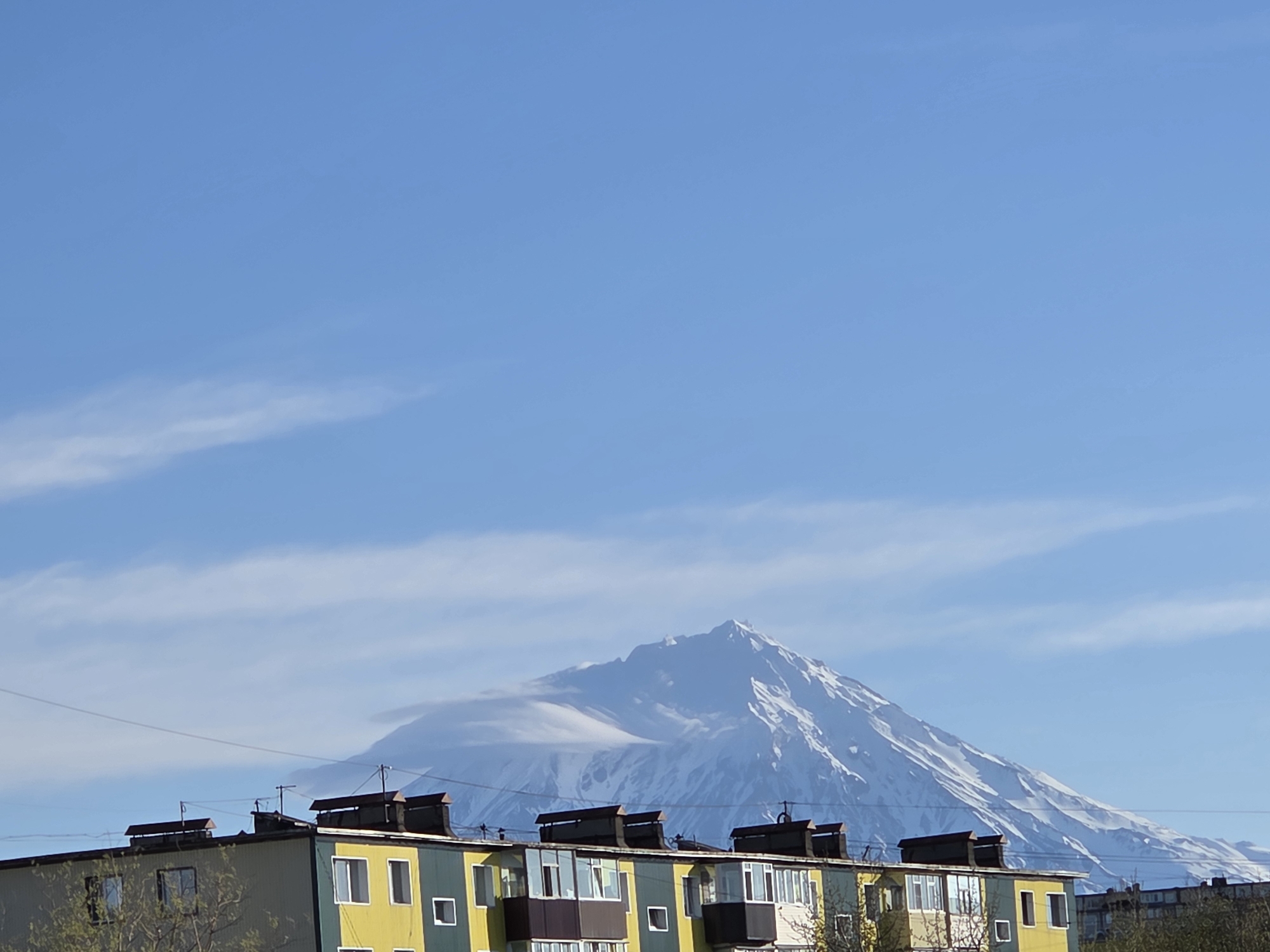 The image depicts a multi-story residential building in the foreground, with a large snow-capped mountain dominating the background. The building has a mix of yellow and green painted walls, with dark-colored chimneys on the roof. Windows are visible on the building's facade. The mountain has a cloud formation partially covering its peak. The sky is blue with sparse, wispy clouds. Power lines are strung across the frame in the midground. The location appears to be in a city with a mountainous backdrop.