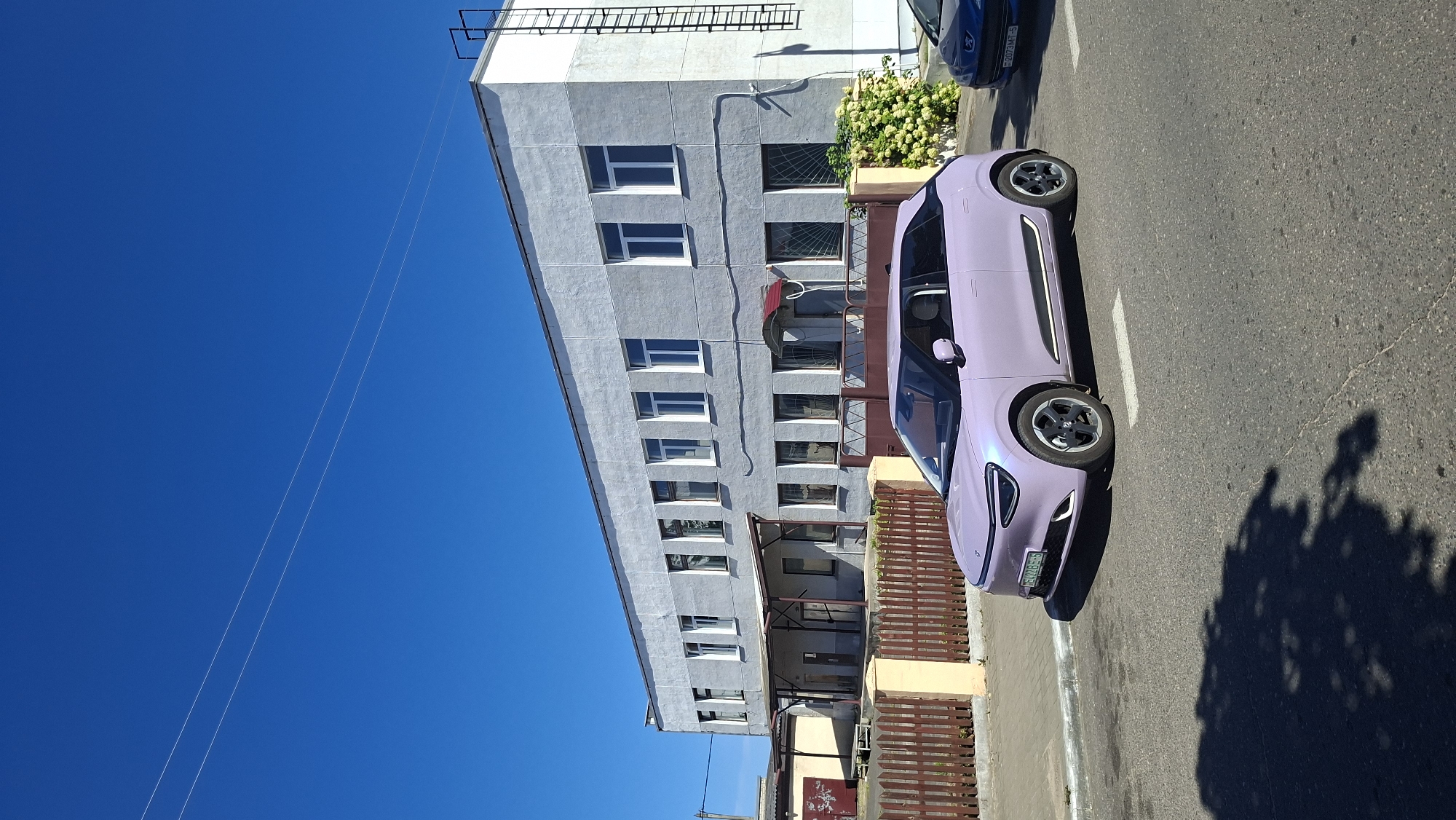 In Myadzyel, Belarus, a light purple electric vehicle is parked on an asphalt road in front of a light gray, two-story building with multiple windows. The building appears to be an industrial or commercial facility, with a fence and a gate separating it from the road. A smaller, yellow building is visible to the left of the main structure. The sky is clear and blue, with power lines stretching across the frame. The car has a license plate that reads "BE2422BE-6". Another blue car is partially visible to the right of the purple car. The scene is illuminated by daylight, casting shadows on the road and the ground.
