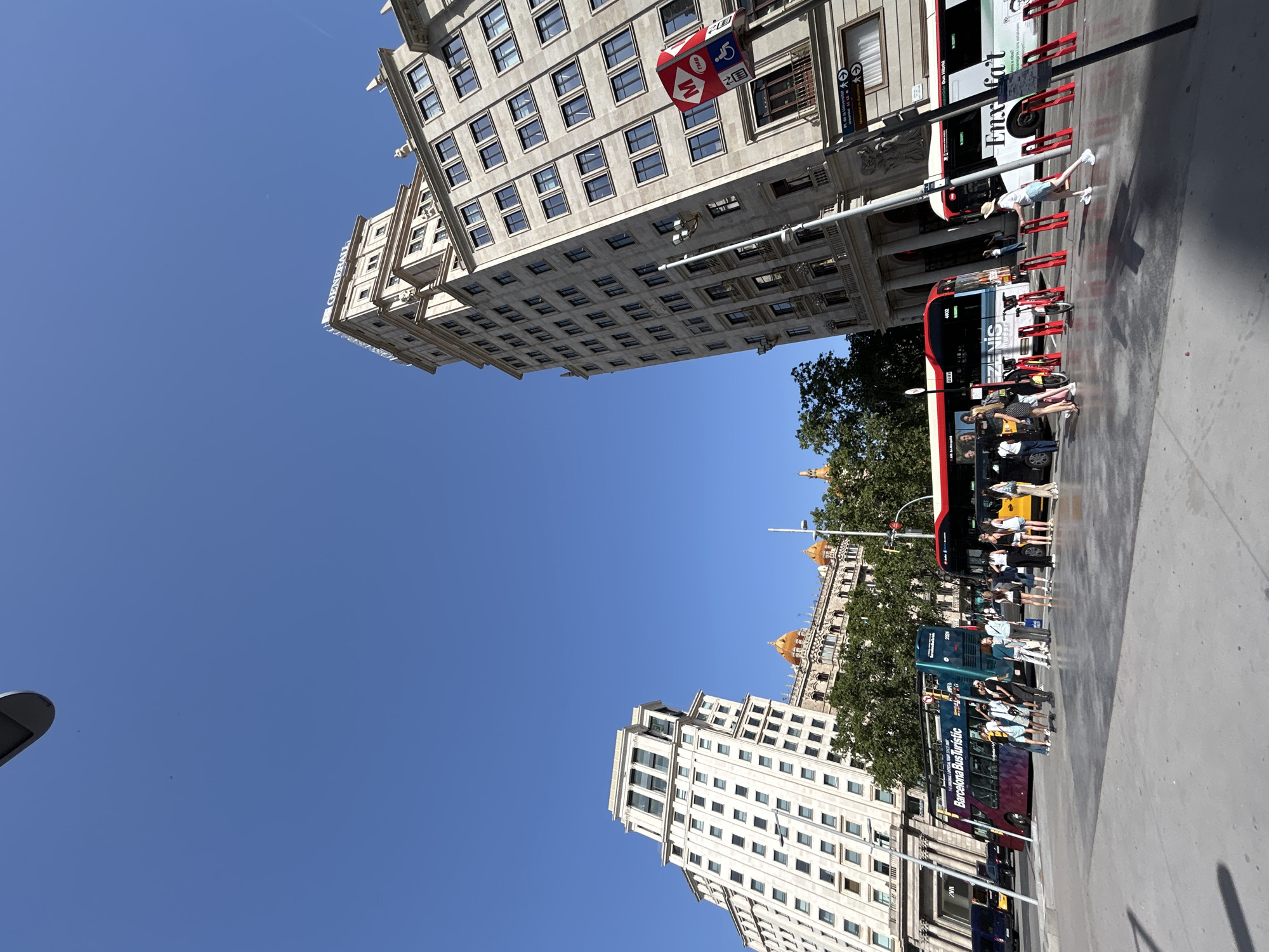This is an outdoor, ground-level, eye-level shot of a wide, modern plaza in Barcelona, Spain, on a clear, sunny day. The plaza is paved with light gray concrete and features a row of bright red bike racks. Several people are standing and walking in the plaza.
In the background, two tall, European-style buildings dominate the skyline. The building on the right is a light beige stone structure with numerous windows and ornate architectural details. The top of this building features the blue text "GENERALI" on a white background. A red and white sign with a "M" logo, indicating a metro station, is affixed to a pole in front of this building. The building on the left is also tall, with a white facade and many windows, and has a tiered, setback design. Behind these buildings, other structures are visible, including a building with orange domed roofs.
Several buses are present in the scene. A double-decker tour bus, colored teal with a purple and pink design, is parked on the left, with the text "BARCELONA BUS TURISTIC" visible. To the right of this bus, a red and white bus is stopped, with people visible boarding or disembarking. Further to the right, another white bus with green and black lettering, including "Enxai'at," is also stopped. Trees line the area behind the buses. In the far left of the image, a dark blue van and a yellow car are visible.
The sky is a clear, vibrant blue, with no clouds. The lighting suggests it is daytime, likely late morning or early afternoon, with the sun casting some shadows on the plaza.