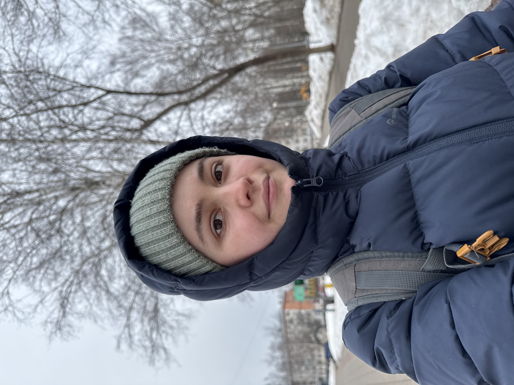 A female subject is centrally framed in a close-up, chest-up view. She wears a dark blue puffer jacket with the hood drawn up, enclosing a light green ribbed knit beanie hat. A grey backpack with a visible orange plastic buckle on the strap is worn over the jacket. Her face is directed towards the camera, displaying a neutral expression. The background reveals a street scene in Izhevsk, Russia, under an overcast sky. Patches of snow and slush cover the ground alongside a paved pedestrian area. Bare deciduous trees are visible throughout the midground and background. Multi-story apartment buildings with faded facades are present on the left side of the frame, and yellow construction equipment is faintly visible in the far right background.