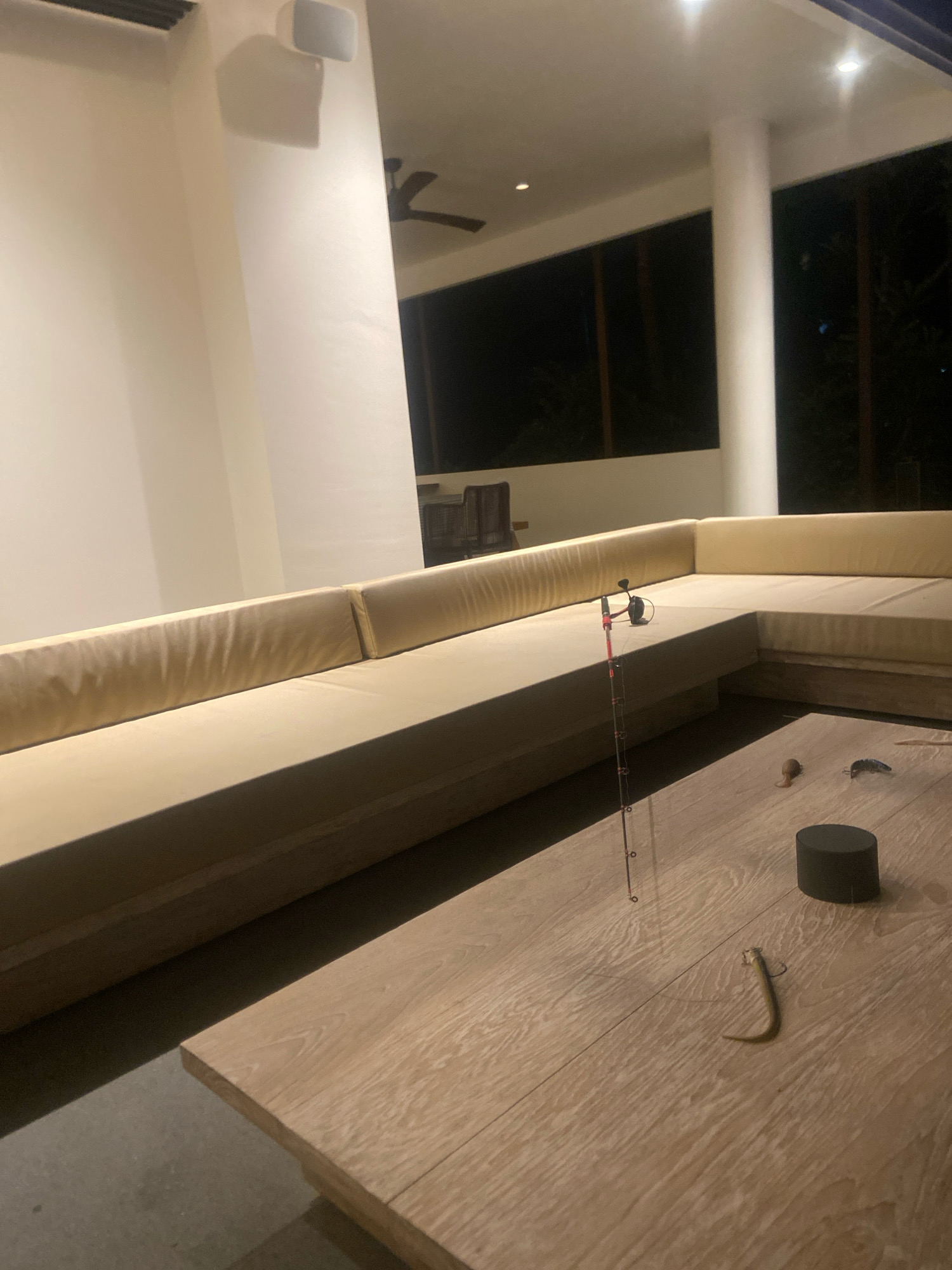 An outdoor lounge area at night features a large L-shaped sectional sofa with light beige cushions. A wooden coffee table sits in front of the sofa, displaying a small fishing rod with a reel, a dark cylindrical object, and two fishing lures. In the background, a ceiling fan is visible on a covered patio, and beyond that, dark vegetation suggests a natural setting. The lighting is dim, with recessed lights on the ceiling illuminating parts of the area. There are no people in the image. The location is a modern lounge in Ngaglik, Indonesia.