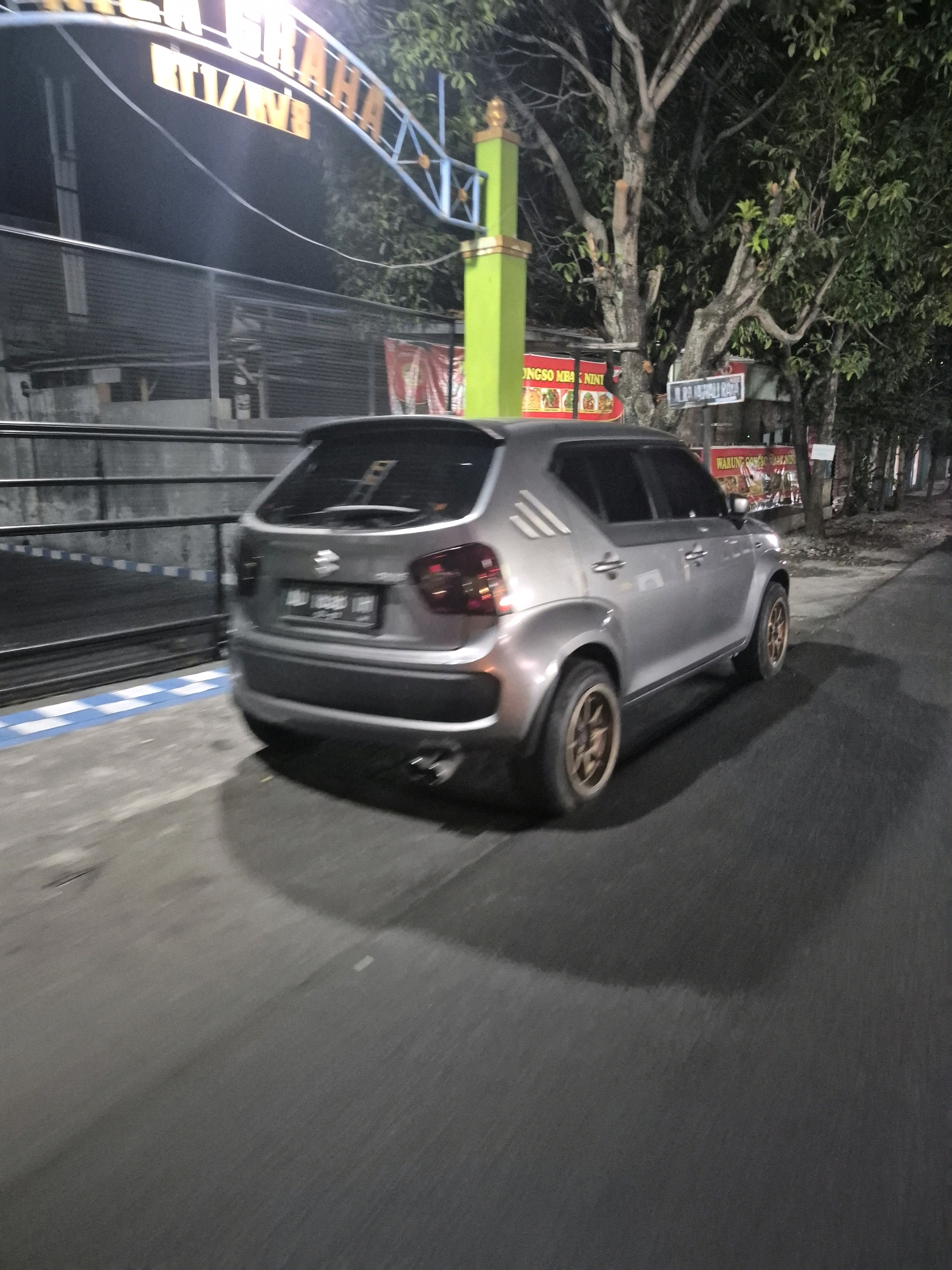 The image is a medium shot of a silver Suzuki Ignis car driving on a street at night in Pabelan, Indonesia. The car is in motion, as indicated by the motion blur. The street is paved and appears to be an urban or suburban road. On the left side of the road, there is a black metal fence with blue and white markings on the ground in front of it. Behind the fence, a building is partially visible.
Above the fence, there is a large, illuminated sign with text in orange and blue. The text on the sign is partially obscured and difficult to read in its entirety, but some words like "RAHA" and "TIZANS" are visible. Further back, there are trees with dense foliage and some streetlights.
On the right side of the street, there are several storefronts with illuminated signs. One sign reads "WARUNG CONGO" with images of food. Another sign says "ONGSO MBA NINI". There are also trees lining this side of the street. The overall lighting suggests it is evening or nighttime.