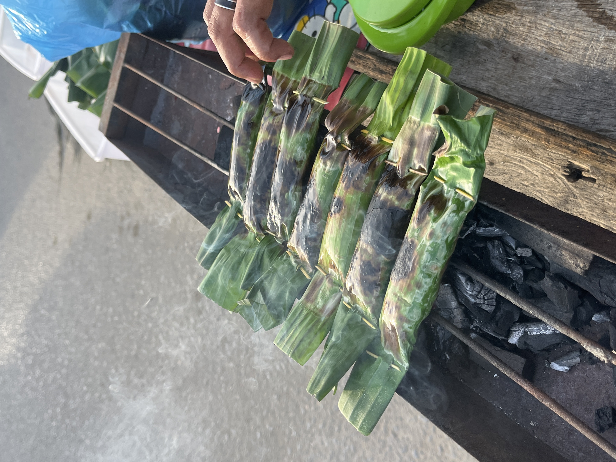 In Lhokseumawe, Indonesia, a person is grilling food wrapped in banana leaves over charcoal. The scene is outdoors, likely a street food stall, with a sandy or concrete ground visible. Several parcels of food, skewered and grilled, are arranged in a row on a metal rack. The banana leaves are charred in places, indicating the cooking process. Smoke is rising from the grill, suggesting the food is actively being cooked. A person's hand, wearing a ring, is visible, seemingly tending to the food. In the background, there's a blue plastic bag and a bright green container. The lighting suggests it's daytime, possibly late afternoon, as the light appears warm. The overall impression is of a casual, local food preparation setting.