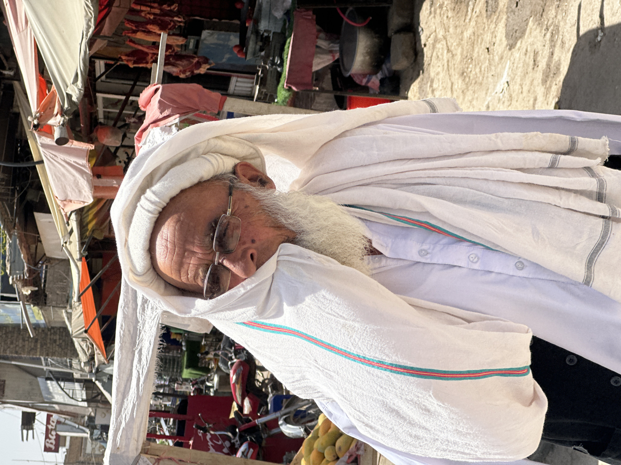 In the city of Kanozai, Pakistan, a man with a long white beard and glasses is pictured. He is wearing a white turban that partially covers his face, and a white button-down shirt. A white cloth with red and green stripes is draped over his shoulder and arm. He is standing in what appears to be a market, with stalls and awnings visible in the background. The ground is dusty and uneven, with debris scattered around. There are motorcycles and other items, possibly goods for sale, also present in the scene. The lighting suggests it is daytime, likely a bright, sunny day.