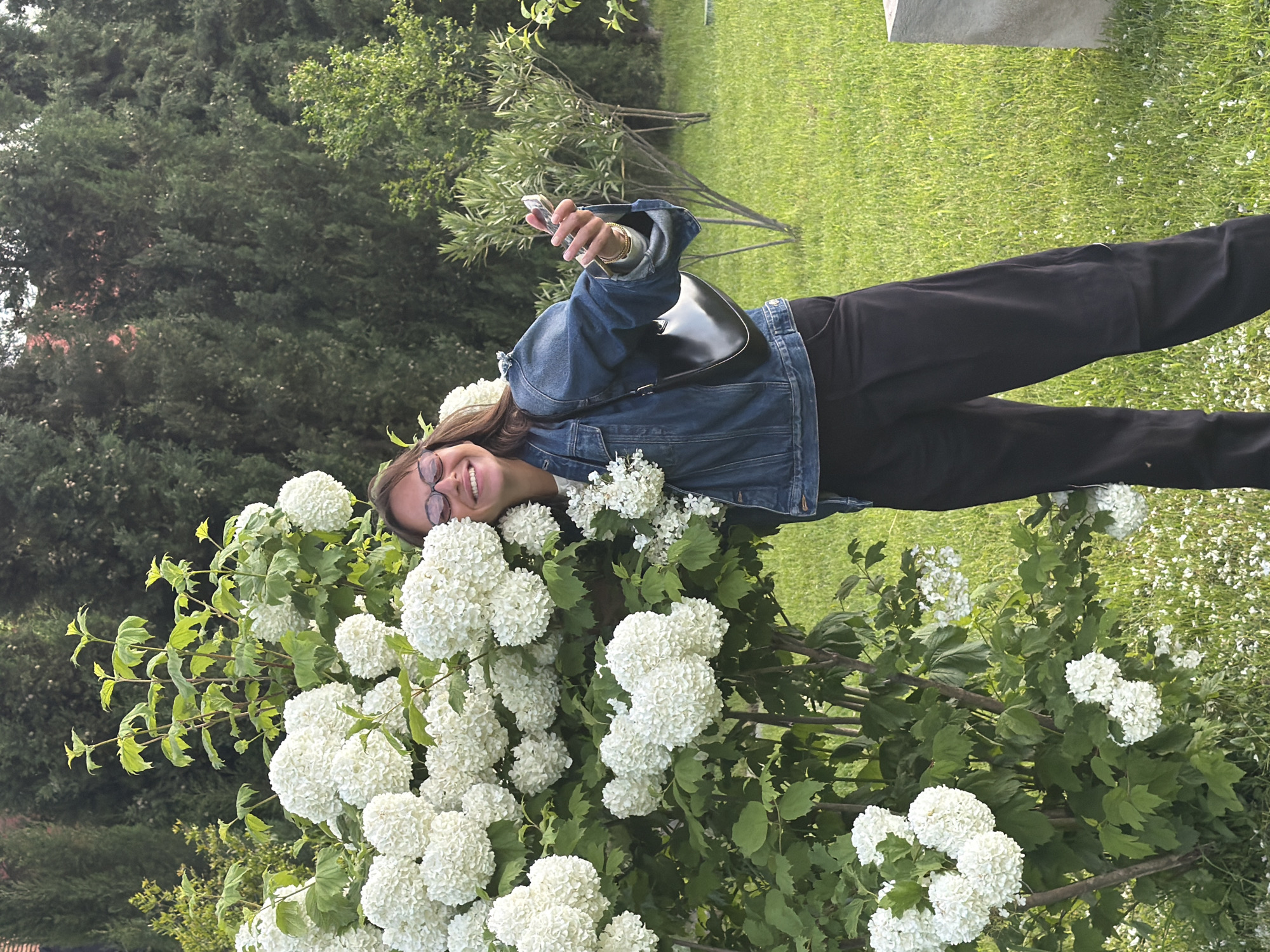 A young woman stands in a garden or park, likely in Napareuli, Georgia, given the prompt context. She poses next to a large flowering bush with numerous white, round flower clusters. She wears glasses, a denim jacket, and dark trousers. A black purse hangs over her shoulder, and she holds a cell phone in her hand. The background includes green grass, trees, and a partially visible concrete structure. The setting is outdoors, with natural lighting.