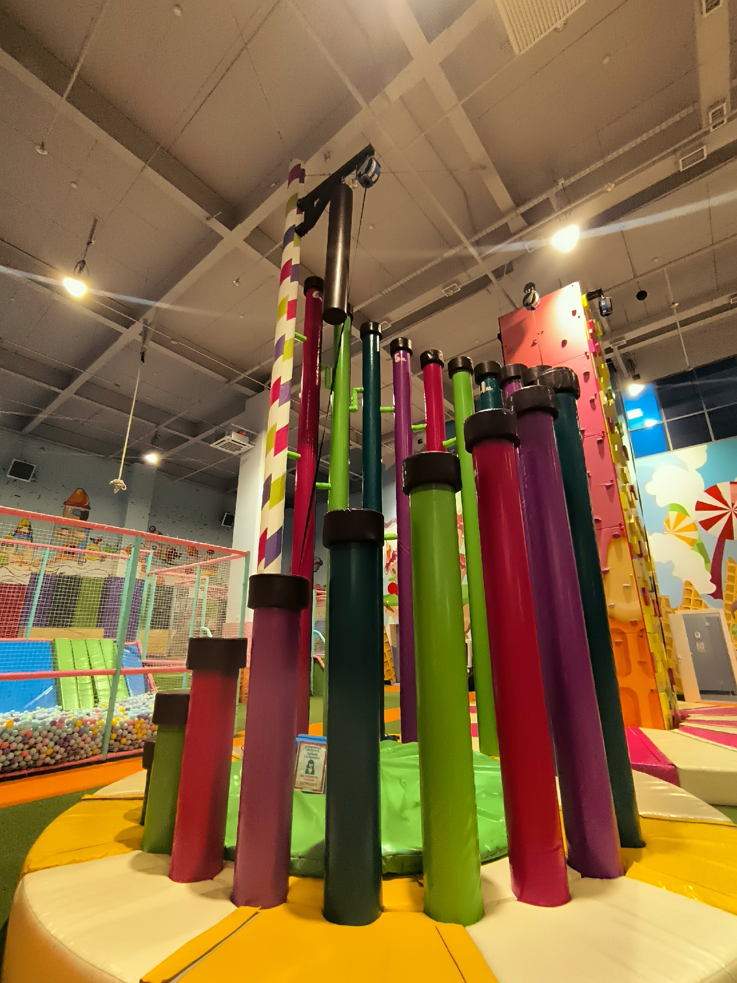 The image shows the interior of an indoor playground. In the foreground, there are several brightly colored, cylindrical obstacles, possibly for climbing or balancing. Behind these, there is a large ball pit filled with multi-colored balls, and a soft play area with netting and colorful padding. In the background, a rock-climbing wall is visible, decorated with a playful design resembling ice cream. The ceiling is a typical drop ceiling with exposed beams and lighting fixtures. The overall impression is a vibrant and active children's entertainment space. There are no people visible in the image. This location is identified as being in the city of Qarağandı city administration, Kazakhstan.