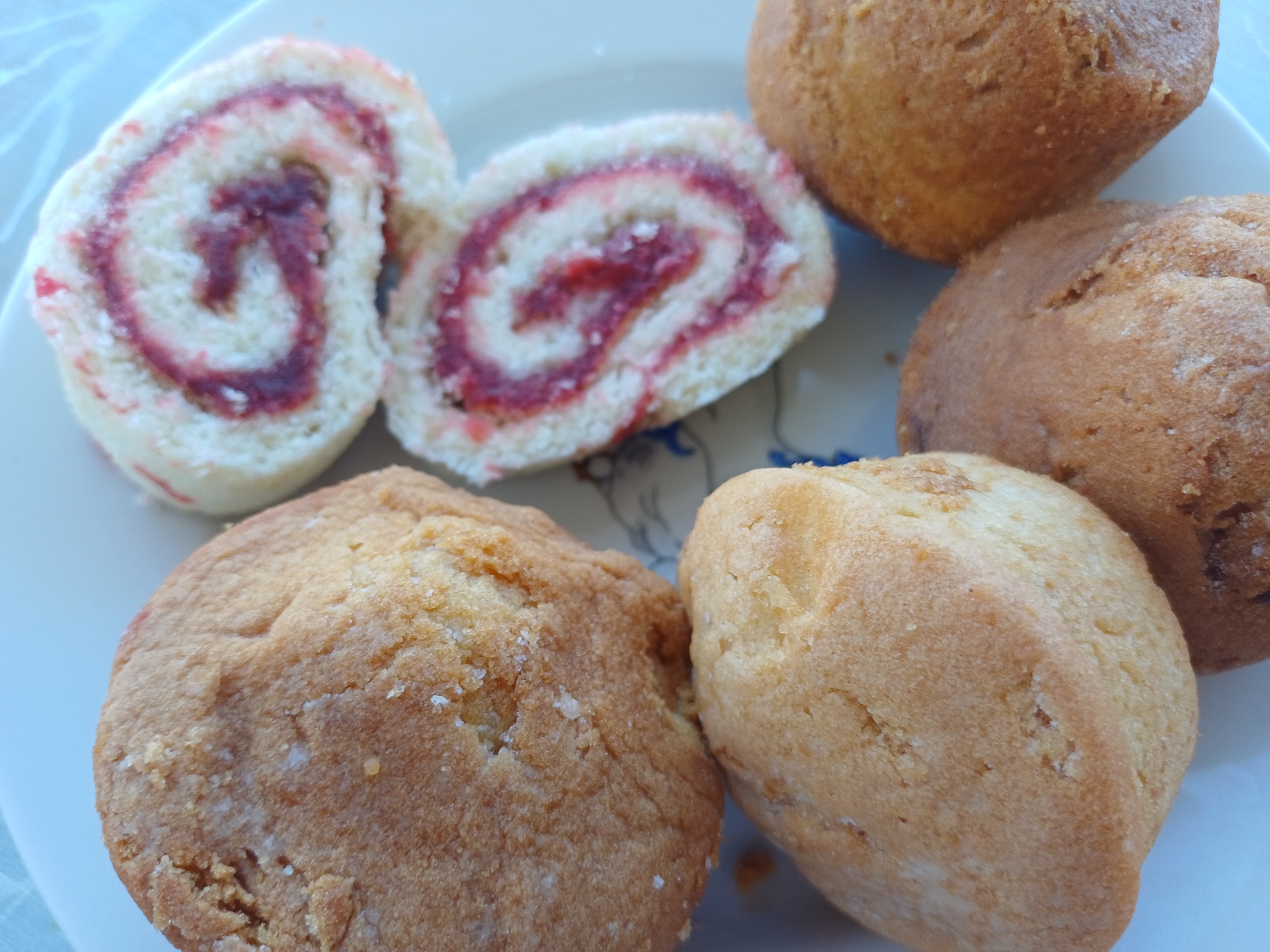 A top-down, close-up shot shows a white plate with several baked goods. The plate is light blue with a faint pattern. In the upper left quadrant of the image is a large, golden-brown, textured cookie. To its right, in the upper right quadrant, are two pinwheel cookies, each sliced to reveal a spiral of white cake and a dark red jelly filling. In the lower half of the image, there are three more baked goods. The largest, a golden-brown cookie, is in the lower left. Below it and slightly to the right is a small, dark brown cake, possibly a muffin top. To the right of that, in the lower right quadrant, is another golden-brown cookie. All the baked goods appear to be on a flat surface. There is no visible indication of the city of Sochi, Russia in the image.
