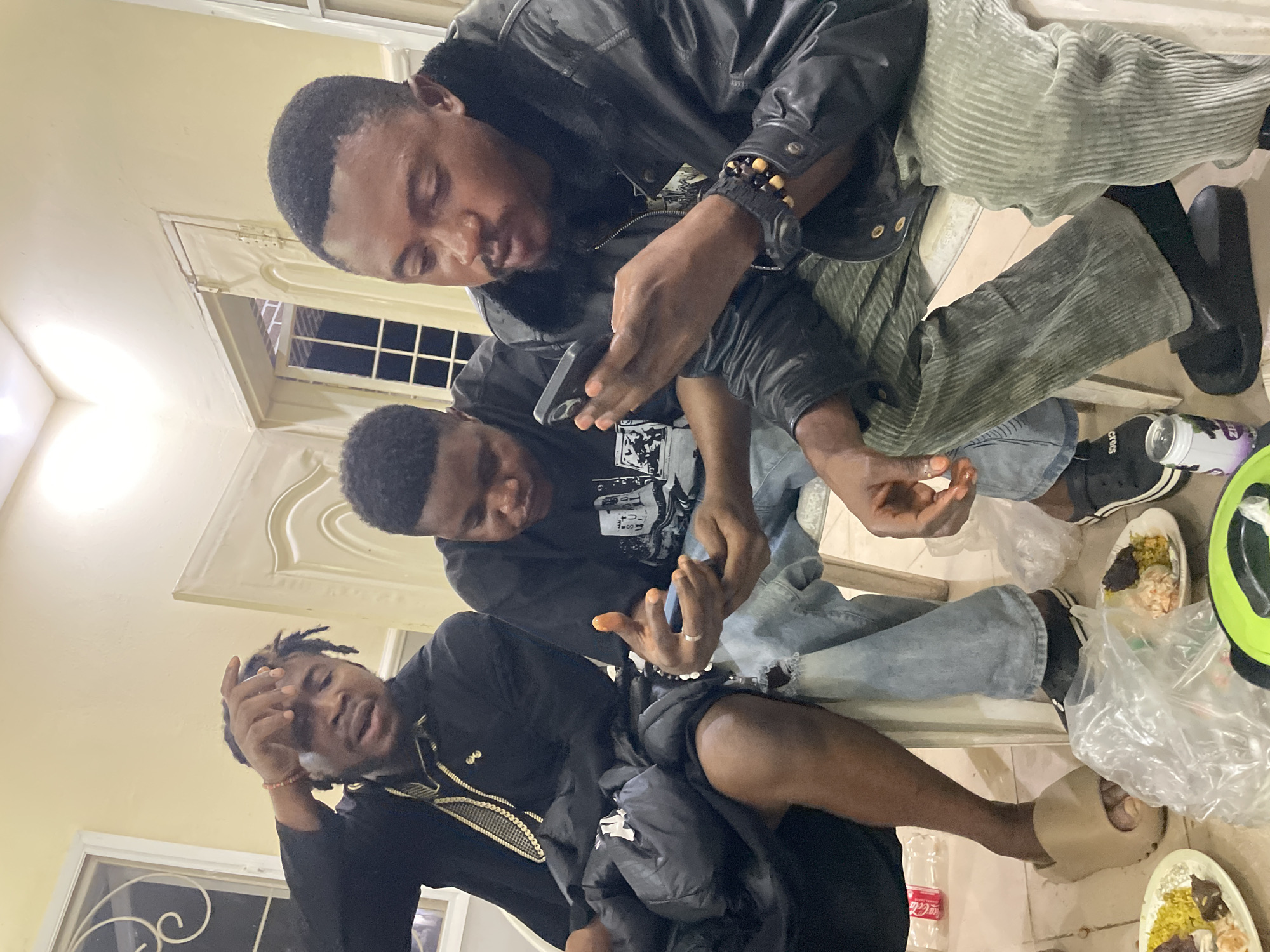 Three young men are sitting close together in an indoor setting, possibly a private residence, though the location in Jos, Nigeria is not evident from the image. One man wears a black leather jacket and greenish corduroy pants. Another wears a black t-shirt and blue jeans. The third man is dressed in a black outfit with gold trim. They are all looking at cell phones. Plates of food are visible near their feet, along with drink containers. The environment appears casually arranged, suggestive of a relaxed social gathering.