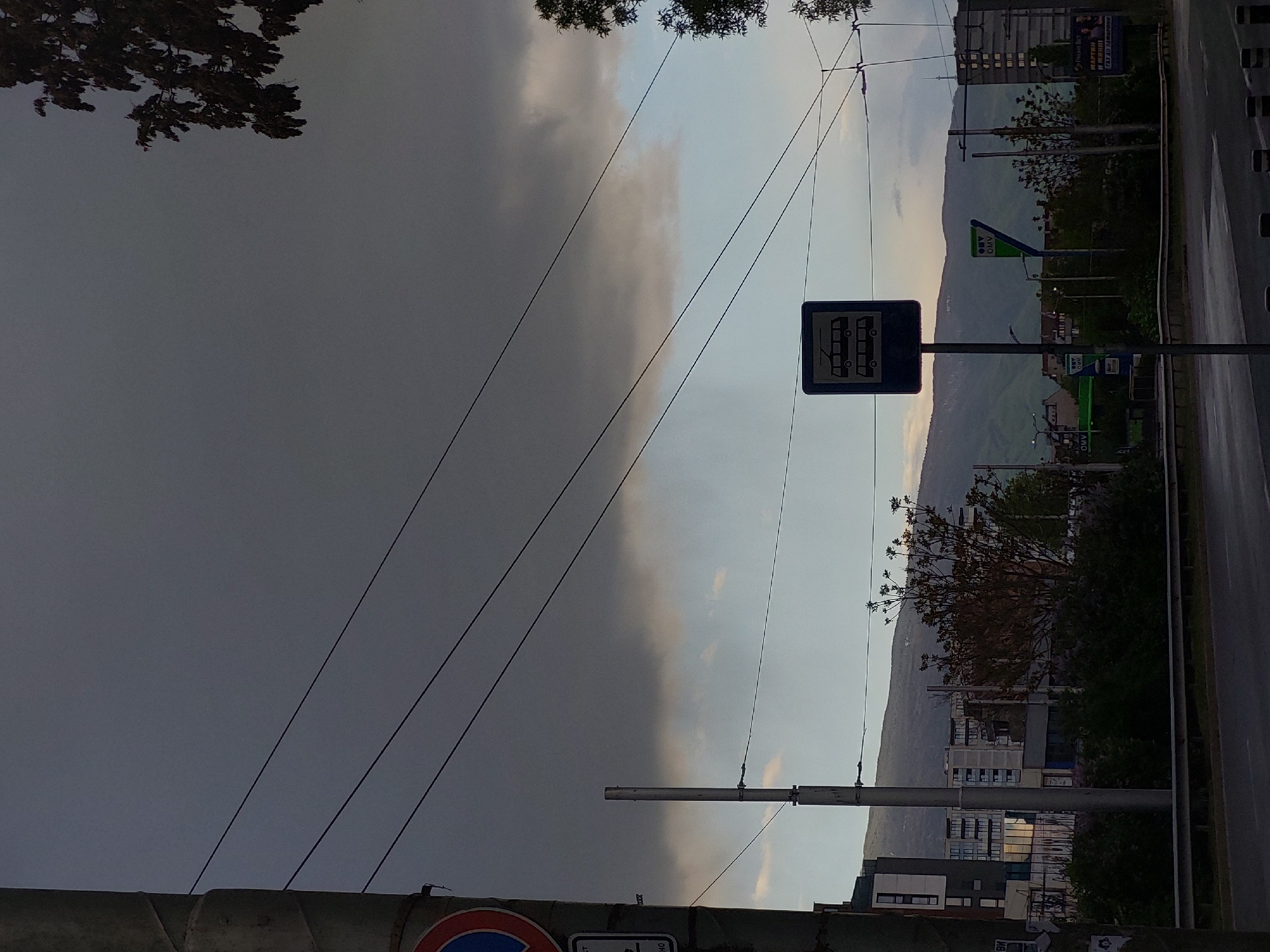 The image depicts a street scene in the city of Unknown, Unknown, featuring a bus stop sign with a trolleybus symbol. The sky is divided between a dark, stormy cloud on the left and a lighter, cloudy sky on the right. Several power lines are visible crossing the sky. In the background, a mountain range can be seen, along with buildings and trees. A road runs along the right side of the frame. An OMV gas station sign is also visible in the background.