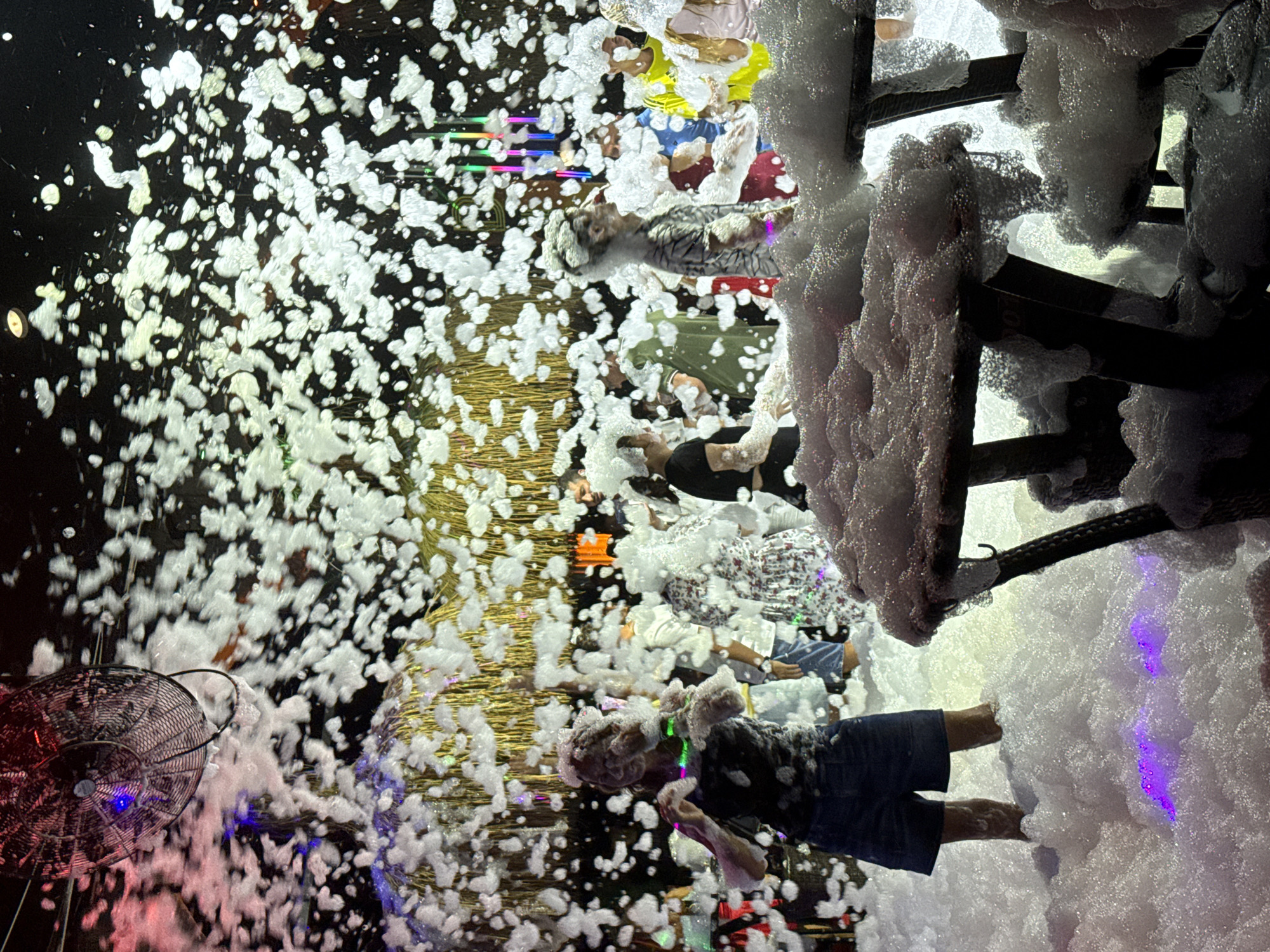 An aerial shot captures a lively scene at a foam party in Alanya, Turkey. The foreground is dominated by two tables covered in a thick layer of white foam. Beyond the tables, a crowd of people are visible, many with foam on their heads, immersed in the energetic atmosphere. Some individuals are seen dancing and interacting, their bodies partially obscured by the swirling foam. In the background, a fan is mounted on the ceiling, presumably contributing to the foam dispersal. The scene is illuminated by colorful lights, casting a vibrant glow on the foam and the revelers, suggesting a nighttime event. The presence of palm-like decorations hints at a tropical or beachside venue.