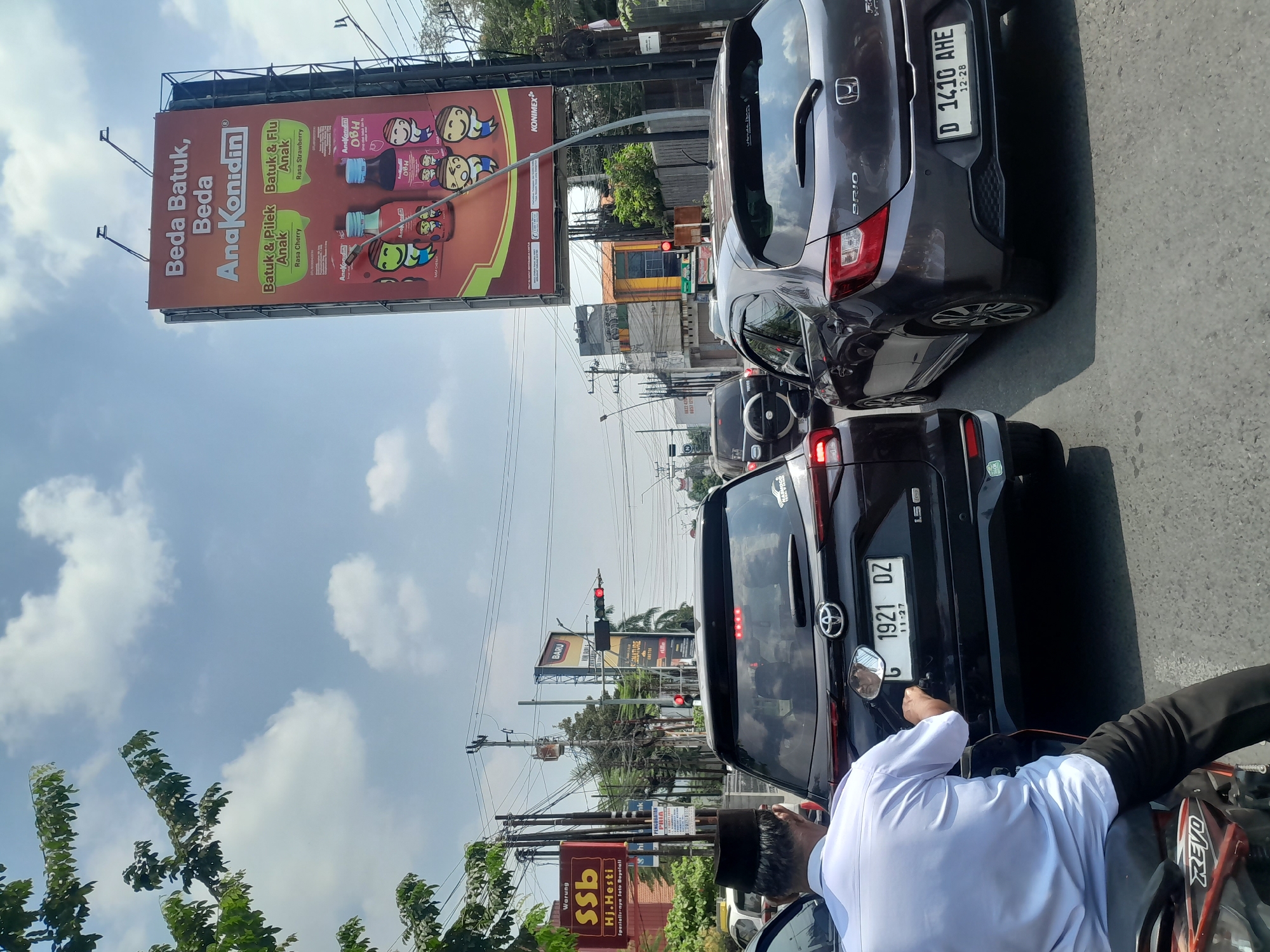 The image captures a street scene in Tegal, Indonesia, likely during daytime due to the bright sunlight and clear sky with scattered clouds. The foreground features the rear of a dark-colored SUV with the license plate "G 1921 DZ" and a motorcycle rider from behind, wearing a white long-sleeved shirt and a black peci hat, on the left. The rider is looking at the SUV's side mirror. To the right of the SUV, a dark gray hatchback with license plate "D 1410 AHE" is visible, facing away from the camera. The road is paved and appears to be congested with traffic, indicated by multiple vehicles and a traffic light in the distance that is red.
Dominating the background is a large billboard for "AnaKonidin," a product for cough and flu, with images of children and product bottles. Below and to the left of this billboard, other signs are visible, including one for "Warung SSB Hj. Hesti" which specializes in Soto Boyolali, and another smaller sign for "BARU SIGNATURE KEKETEK". Electrical poles and wires crisscross the sky, typical of urban infrastructure. Green foliage from trees is present on the left and right edges of the frame, suggesting the presence of roadside vegetation. The overall impression is that of a busy city intersection in Tegal, Indonesia, experiencing moderate traffic.