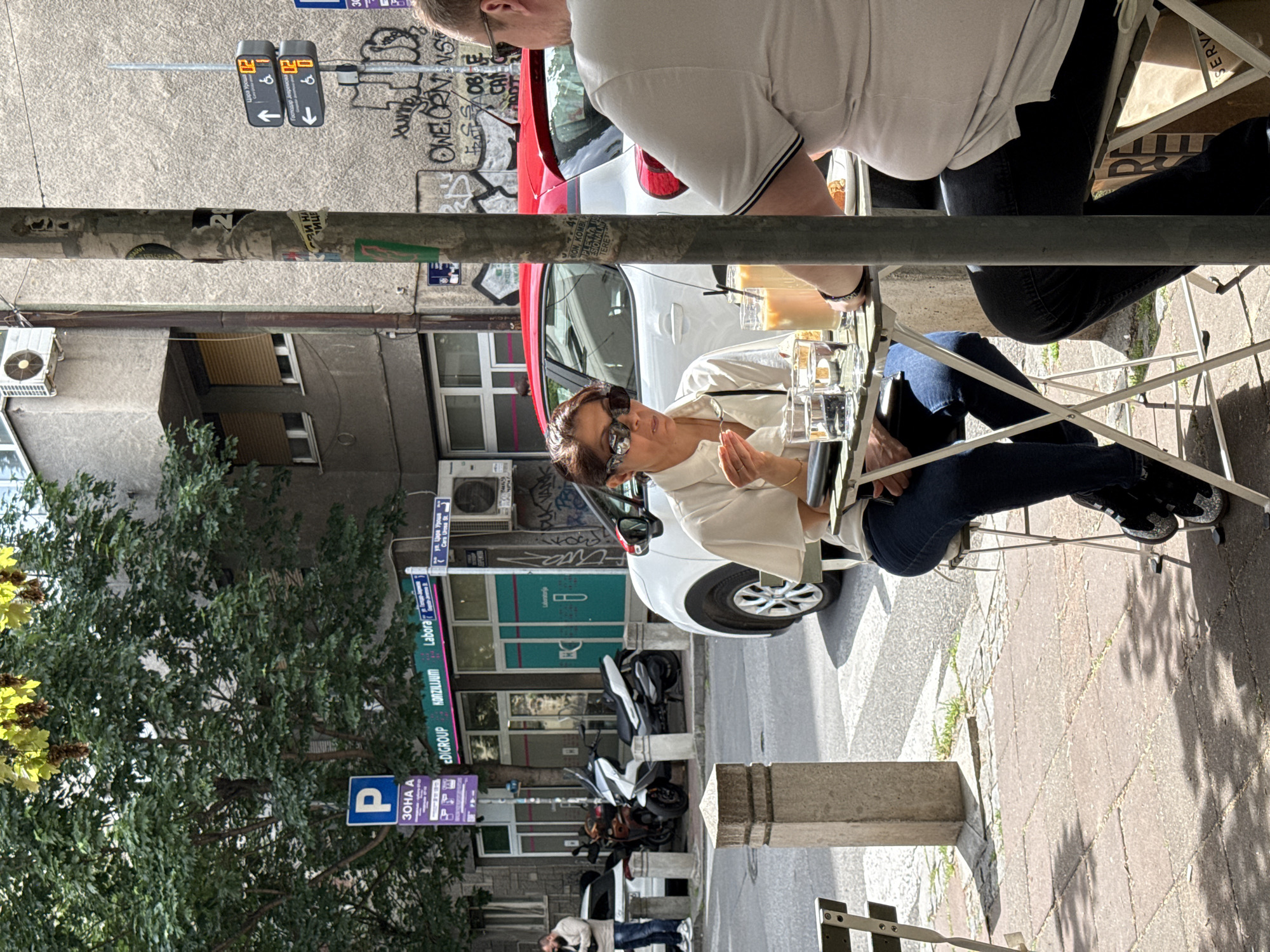 The scene captures a street cafe setting, likely in Belgrade, Serbia, as suggested by Serbian text on nearby signs. A woman in sunglasses and a white shirt sits at an outdoor table, interacting with another person who is partially visible and wearing a white shirt. Several drinks sit on the table.
Behind them, street signs indicate "Цара Уроша," which translates to "Tsar Uroš Street." A parking sign reads "ZONA A." Various parked vehicles, including a white car with red accents and some scooters, line the street. The building facades include commercial signs for "Labora," "EDIGROUP," and "KONZILIJUM." The street is paved, and the overall environment suggests an urban setting. The shadows indicate a sunny day.
