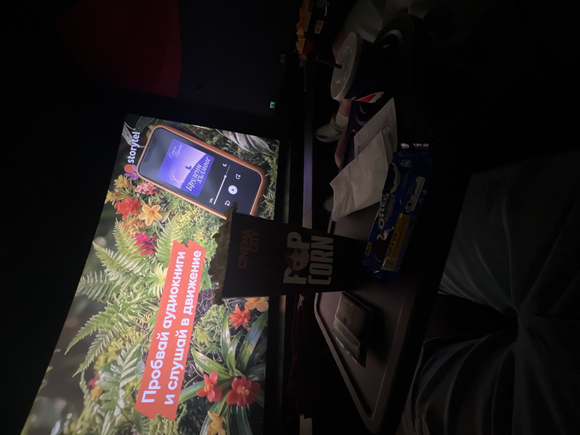 The image shows a scene inside a movie theatre in Sofia, Bulgaria. A patron is sitting in a darkened auditorium, indicated by the large screen in front, where an advertisement for Storytel audiobooks is displayed. The audiobook cover visible on the phone screen features the title "Брулени хълмове" (Wuthering Heights) by Emily Brontë in Bulgarian.
On the tray table in front of the viewer, there's a container of popcorn from Cinema City, a package of Oreo cookies, napkins, and a cup. The viewer's clothing is visible on the right, suggesting they are wearing a light-colored shirt or t-shirt. The overall environment is dim, typical of a movie theatre setting.