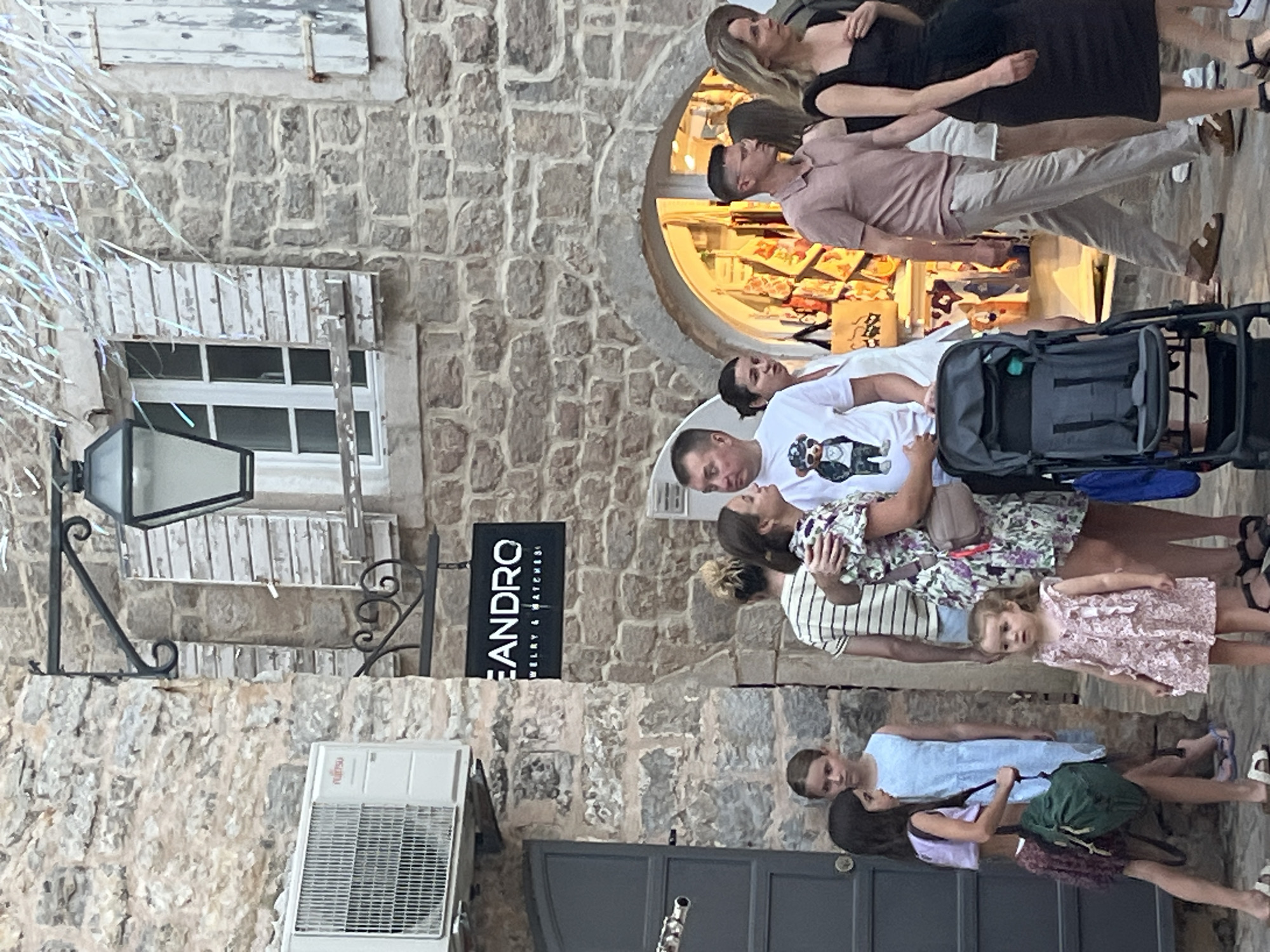 In Budva, Montenegro, a group of people are walking along a stone-paved street. The architecture features rough stone walls and shuttered windows. An air conditioning unit is mounted on the wall to the left, and a black sign that reads "LEANDRO JEWELRY & WATCHES" hangs above a doorway.
In the foreground, a man in a white t-shirt with a bear graphic embraces a woman in a floral dress. A stroller is parked beside them. To their left, two young girls stand together, one in a light blue dress and the other in a purple top and patterned skirt, carrying a green backpack. A toddler in a pink dress stands to the right of the stroller, looking towards the left. Further back, a man in a pink polo shirt and light pants walks with a woman in a black dress. Other individuals are visible in the background, suggesting a lively street scene. The lighting suggests it is daytime, possibly late afternoon or early evening.