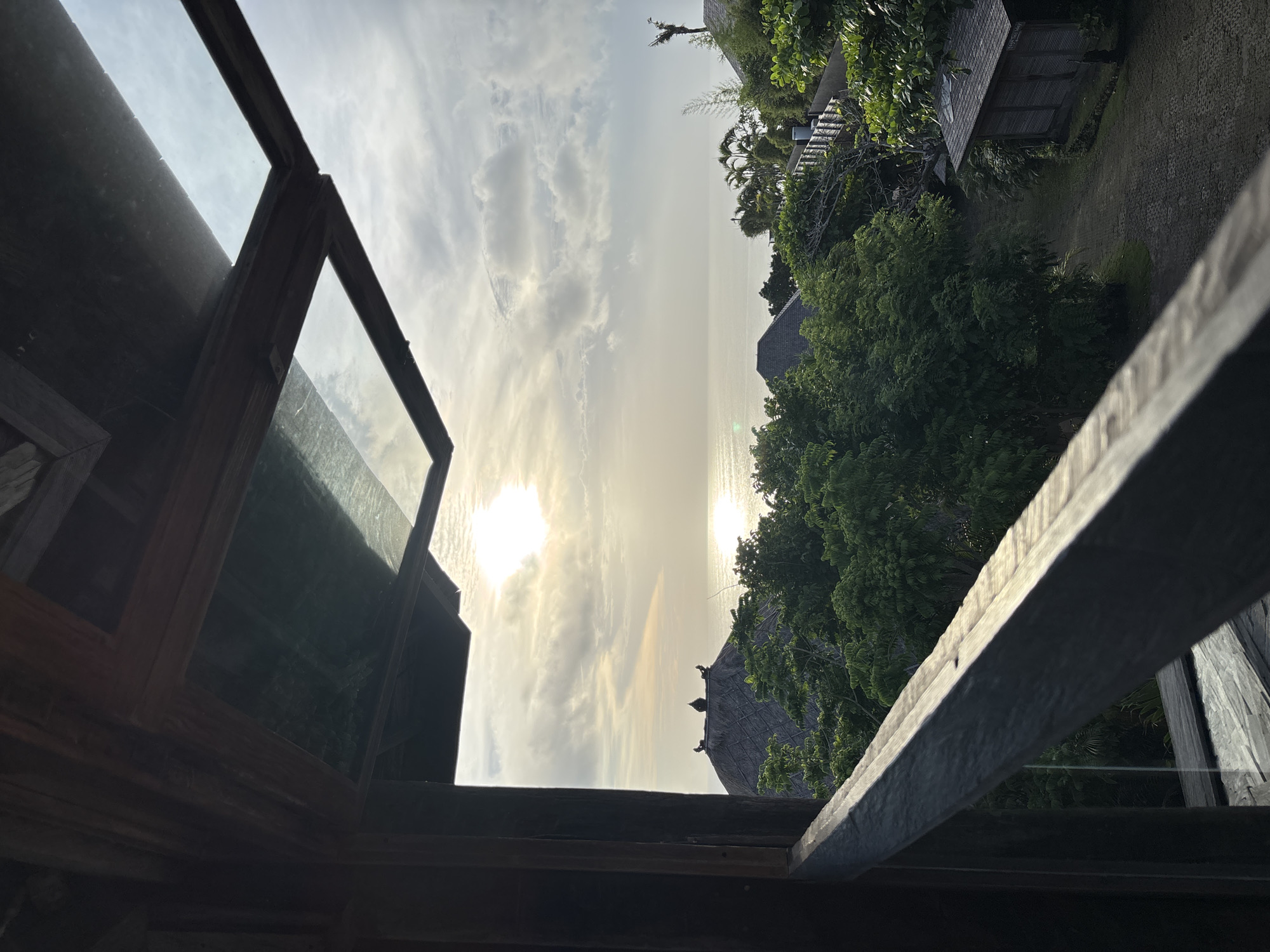 The image shows a view from what appears to be an interior space, through an open wooden window or framework, to an outdoor scene. The foreground is dominated by the dark, weathered wood of the structure, obscuring portions of the view. Beyond the window, the scene is divided into two distinct sections: the sky and a lush, tropical landscape.
The sky is bright, with diffused sunlight shining through clouds. A hint of what seems to be water is visible, possibly the ocean, reflecting the sunlight.
The landscape consists of dense green vegetation, possibly trees and shrubs. Some structures with dark, peaked roofs can be discerned amidst the greenery. A walkway or paved area is partially visible on the right side of the frame, suggesting the presence of buildings or a resort area.
Based on the tropical vegetation and the suggestion of an ocean view, the location is likely Kuta Selatan, Indonesia. The image does not contain any people or actions.