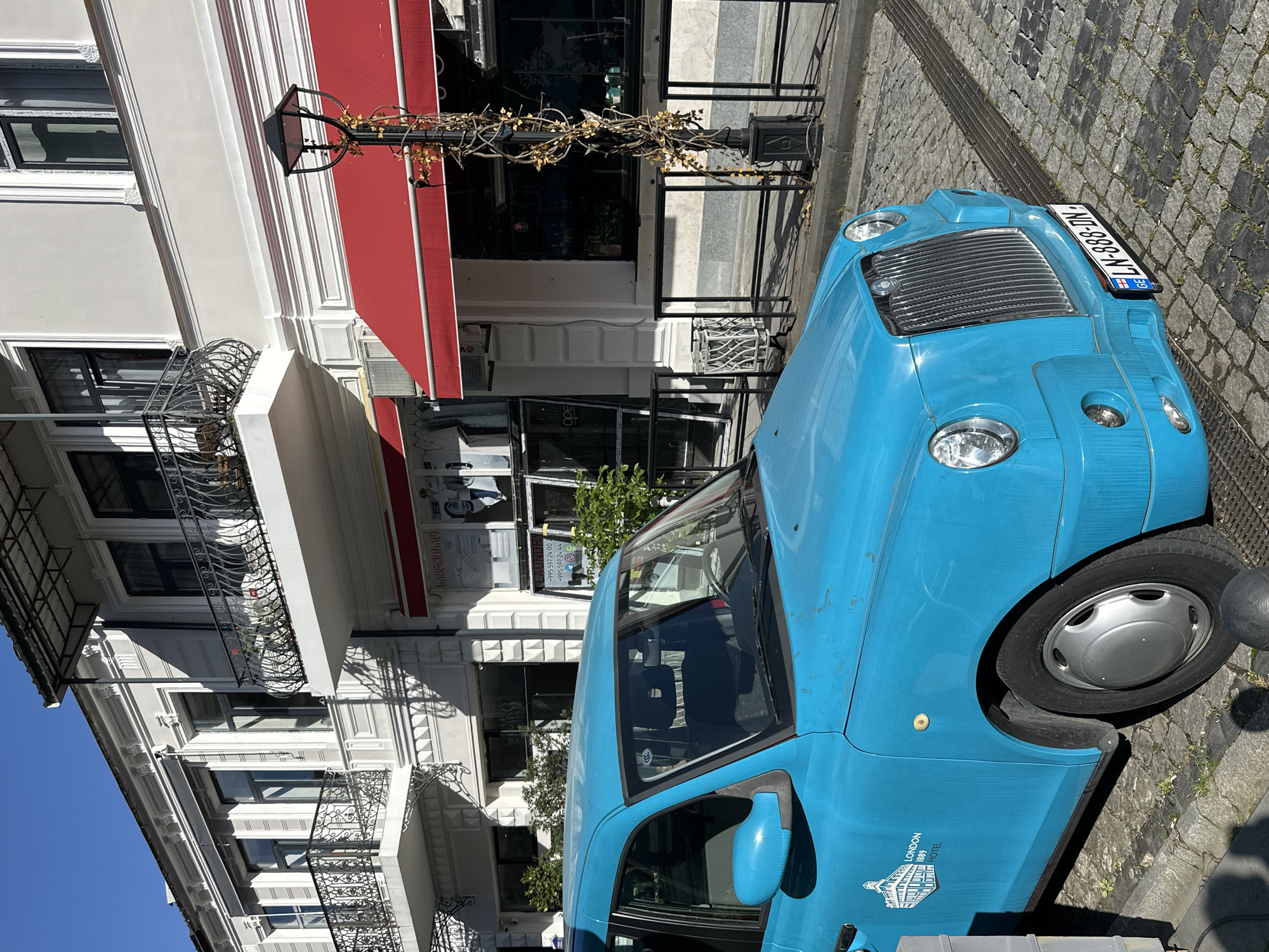 A light blue London-style taxi cab with the Georgian license plate "LN-888-DN" is parked on a cobblestone street in what appears to be Batumi, Georgia. The car features the words "LONDON 1888 HOTEL" and laundry service contact information on its side. The taxi is situated next to a building with a red awning and ornate balconies. The building's storefront has a glass facade with "ορε" visible in the window. A lamppost with decorative vines stands between the building and the car. The background includes architectural details of the building and a glimpse of the sunny sky.