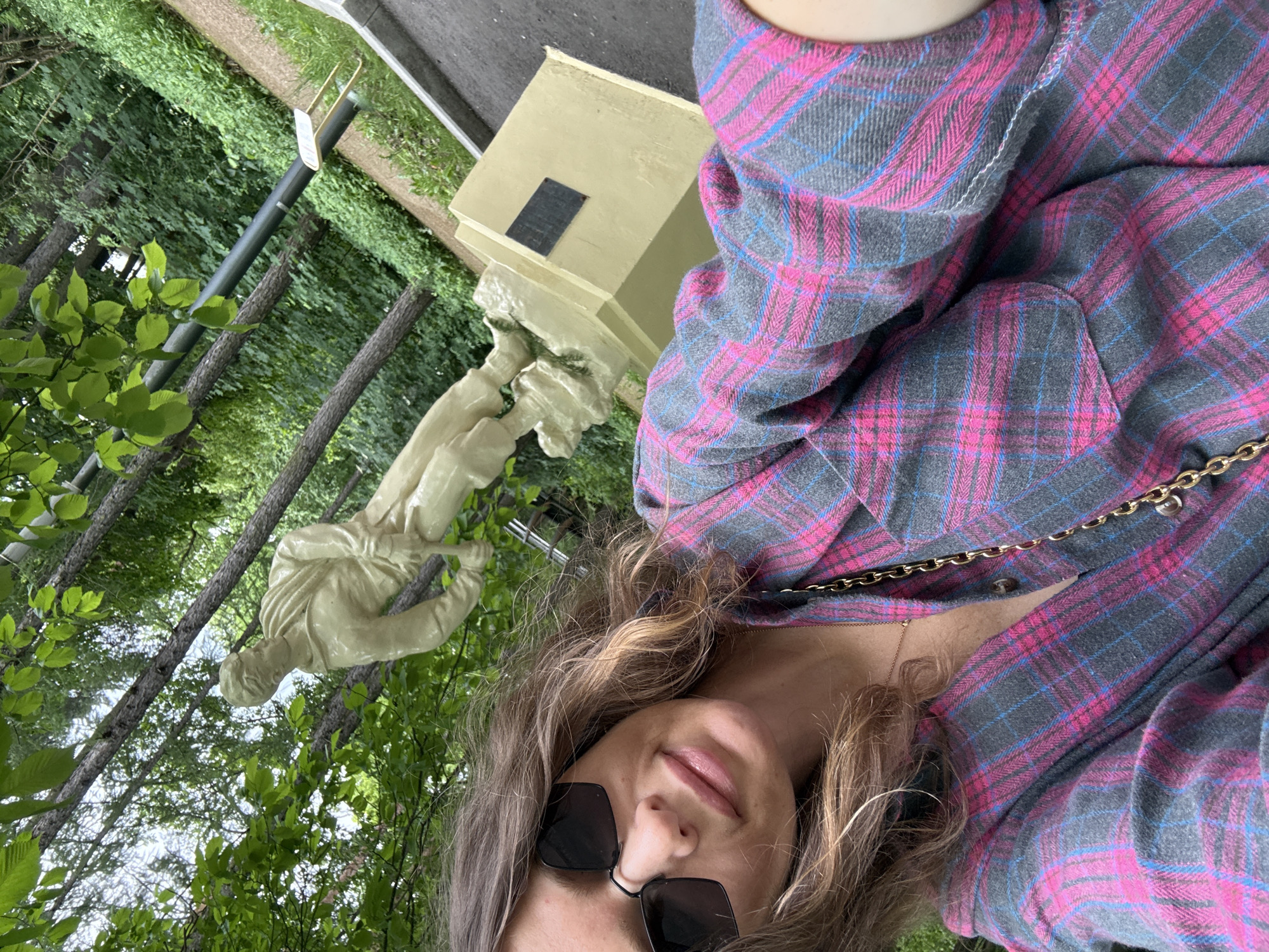 In an outdoor setting, a young woman with light brown hair and sunglasses takes a selfie. She is wearing a pink and grey plaid shirt and a gold chain necklace. In the background, a light-colored statue of a man in a hat and uniform stands on a pedestal. Trees and greenery surround the statue, suggesting a park or public space. The statue appears to be located in Russia.