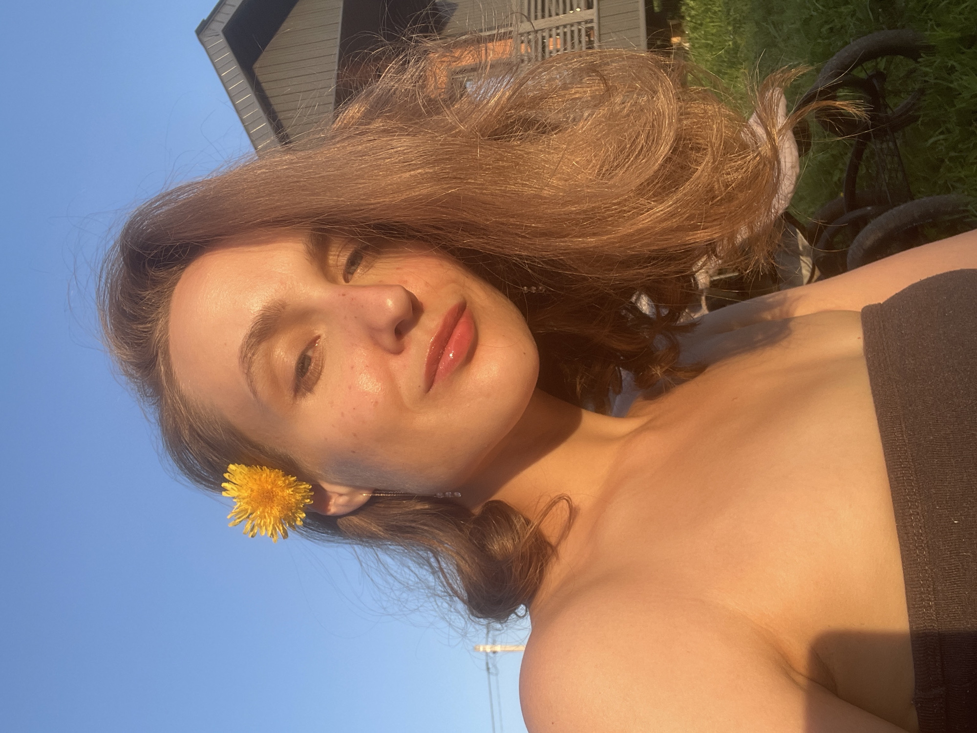 A young woman with shoulder-length brown hair is outdoors in Подшивалово, Russia. She has a dandelion tucked behind her ear. She is fair-skinned with freckles and is wearing a dark tube top. A building is visible in the background along with some greenery and what appears to be a black cart or stroller. The sky is clear and blue.