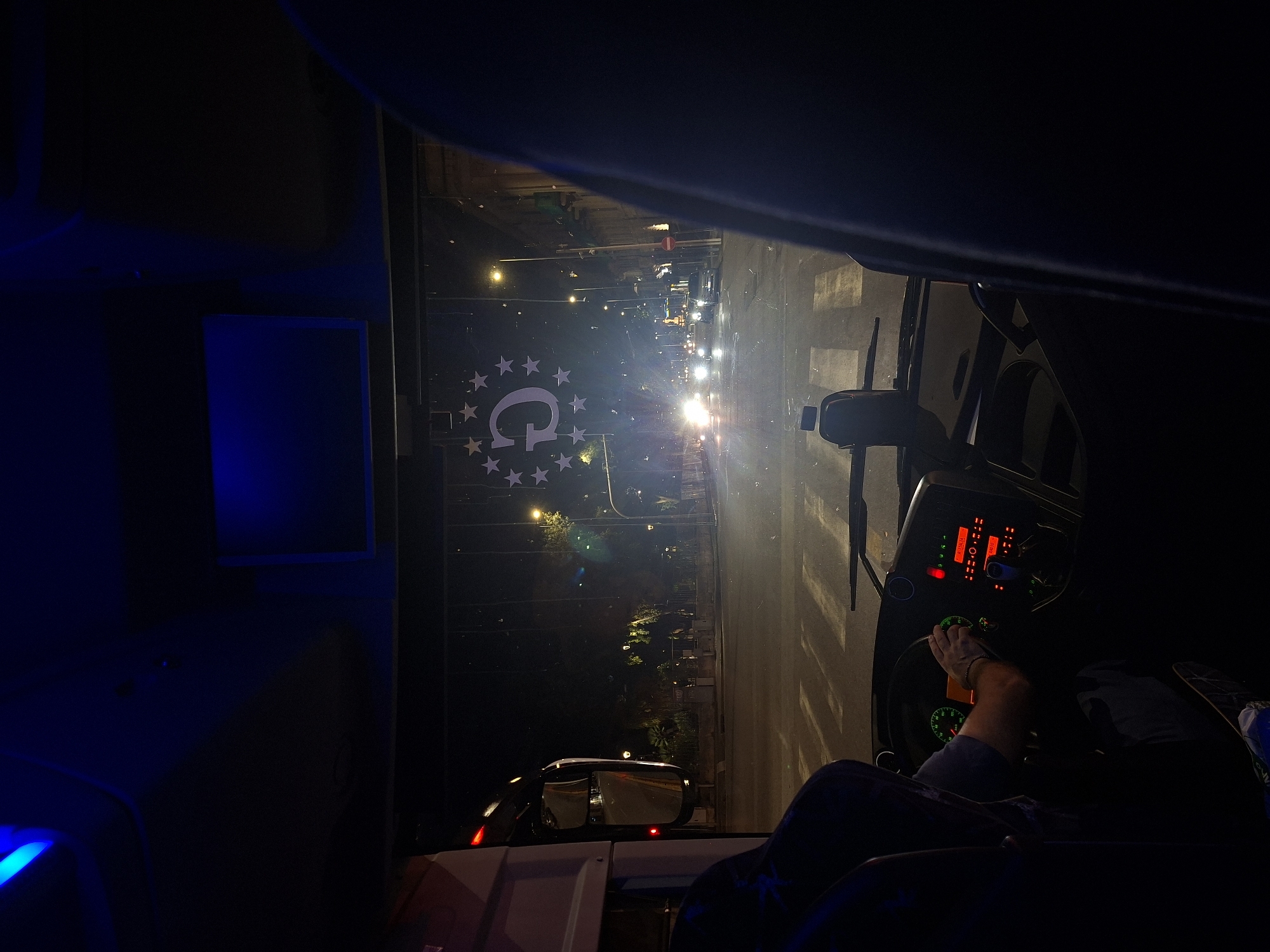 The image depicts a night scene viewed from inside the cab of a vehicle, likely a truck, situated in the city of Groningen, Netherlands. A large "G" logo surrounded by stars is painted on the road surface outside. The driver, whose arm and leg are visible, is holding the steering wheel. The dashboard glows with red lights. Outside the windshield, street lights illuminate a road with pedestrian crosswalks and sparse traffic. Buildings and trees line the street. The vehicle's side mirrors reflect the road behind. The interior of the cab is dark, with a blue light source present on the left.
