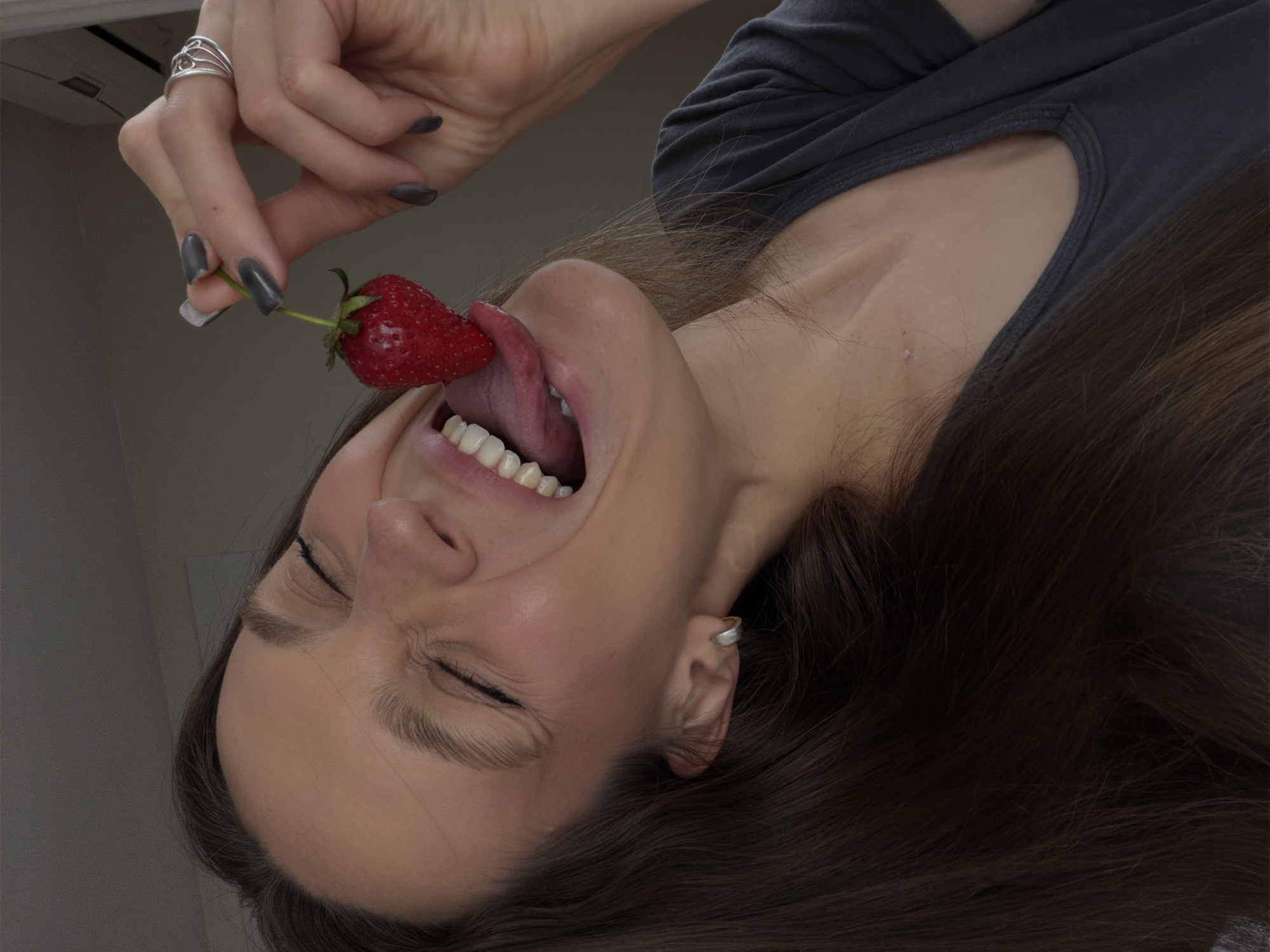 A woman with brown hair and wearing a gray top is holding a strawberry to her mouth. Her eyes are closed, and her tongue is touching the strawberry. She is wearing a ring on her finger and a stud earring. The location is not provided in the image and is therefore unknown.