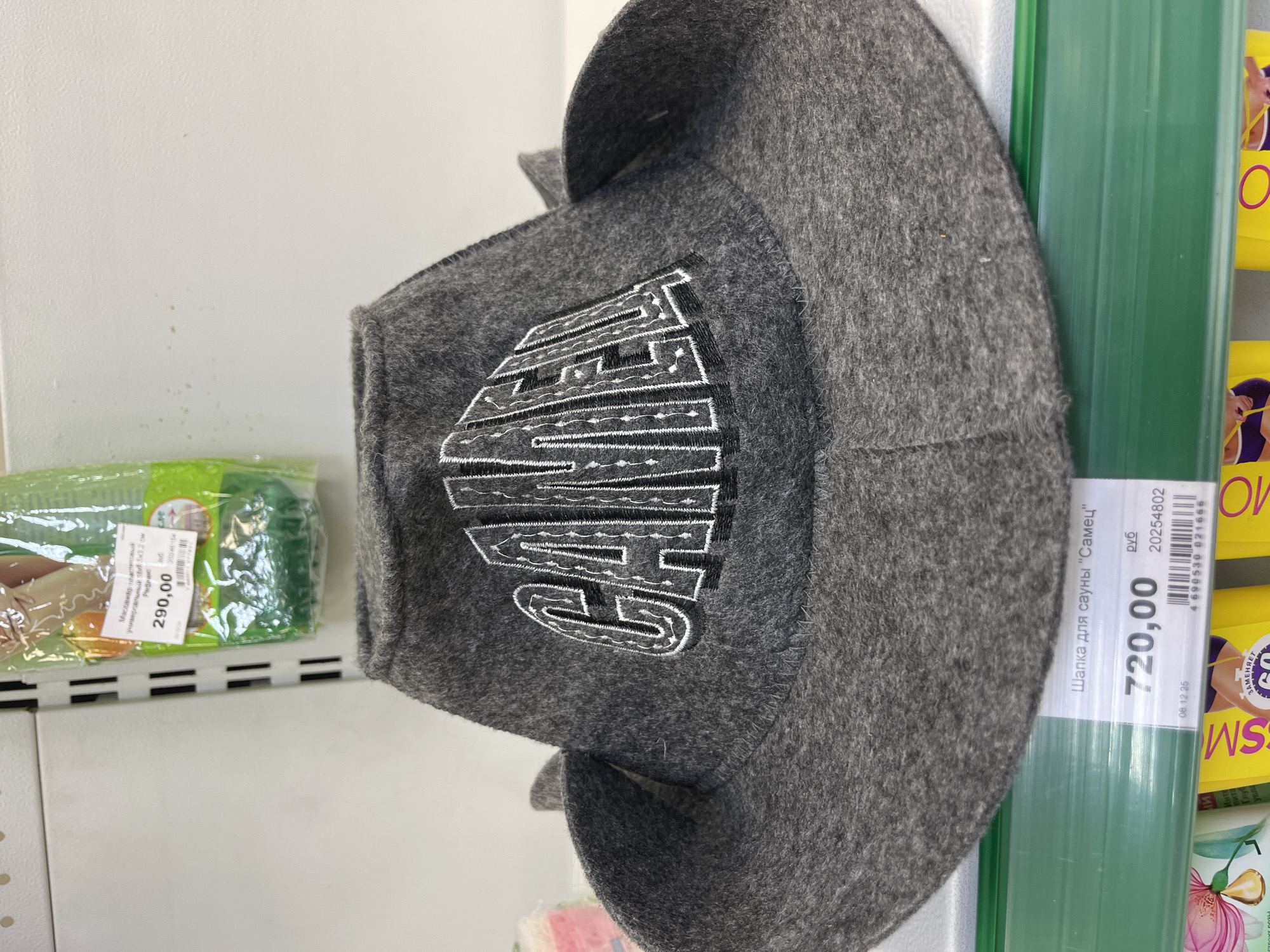 A grey felt hat with the word "САМЕЦ" embroidered in white and black lettering is displayed on a shelf in a store. The hat is positioned centrally in the frame, with its brim spread wide. Below the hat, a price tag is visible, reading "720,00 руб" and a barcode. To the left of the hat, a green shelf is visible, and on the far left of the image, a pink object with a floral design is partially visible. To the right of the hat, the background is a plain white wall. The image appears to be taken in a retail environment.