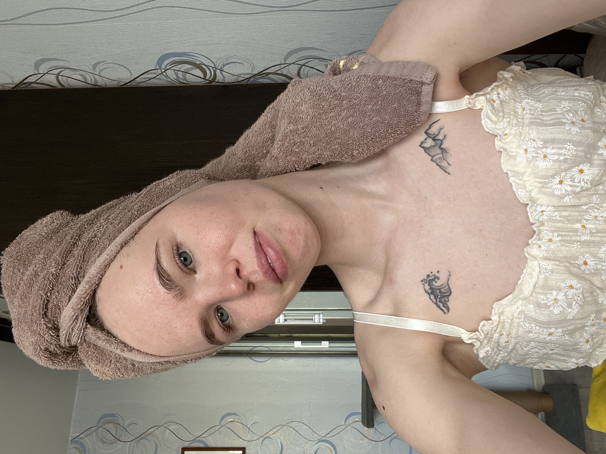 A rotated close-up shot of a woman with light skin and blue eyes. She has a brown towel wrapped around her head. She is wearing a light-colored top with daisy patterns. Two small tattoos are visible on her chest/collarbone area. The background includes wallpaper with a wavy pattern and a dark brown door or panel. The location cannot be determined from the image with certainty, but the presence of the city of Chaikovsky, Russia, cannot be confirmed.