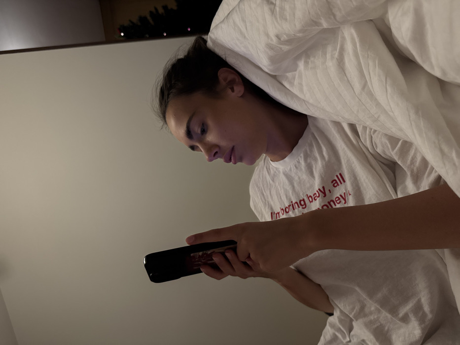 A young woman lies in bed looking at a phone. She is partially under white covers and wearing a white t-shirt with red lettering. The text on the shirt reads "I'm boring buy, all money." Her hair is pulled back. A dark horizontal shape, possibly a picture frame or shelf, is visible behind her head. The wall is a light color.
