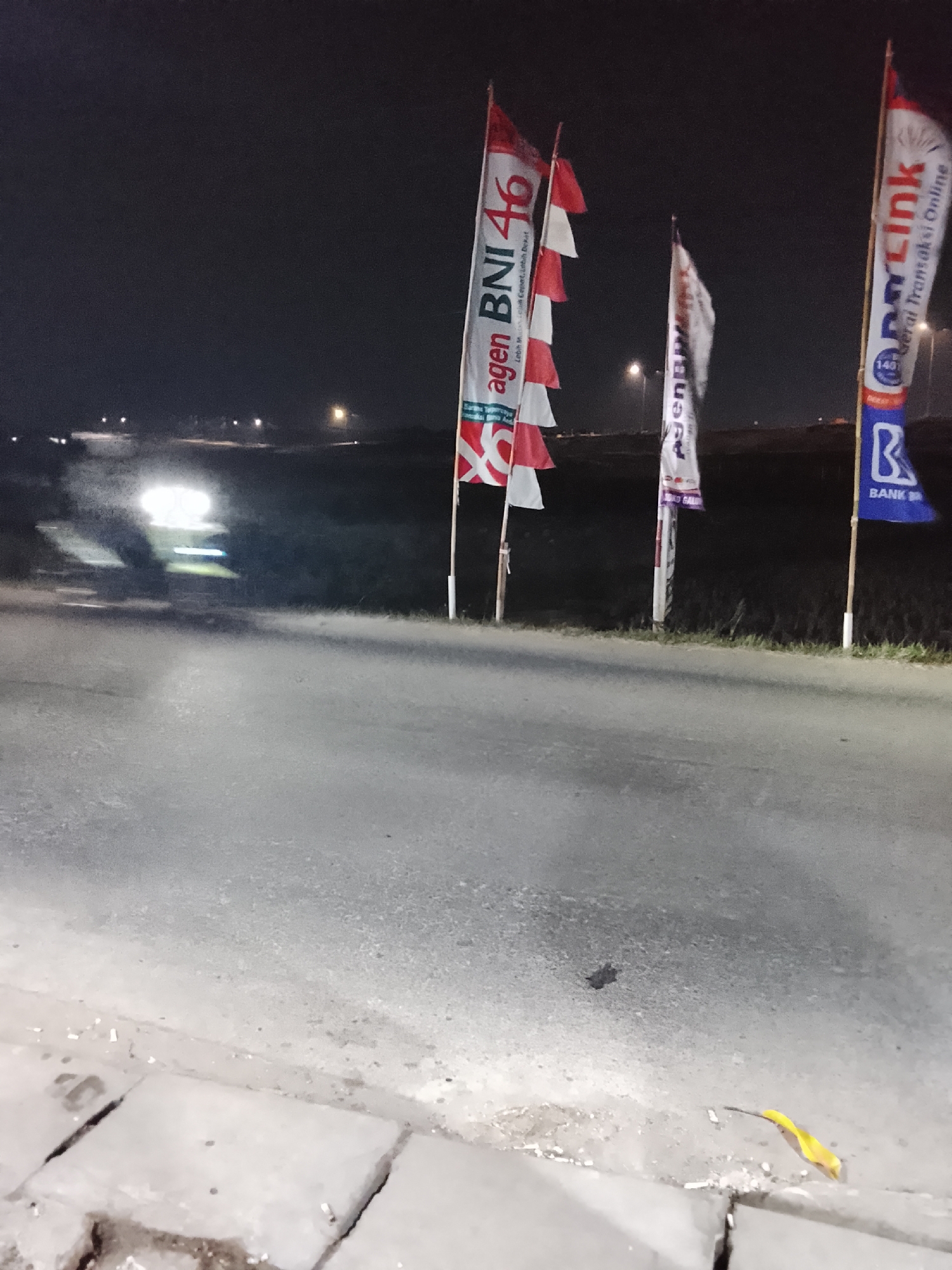 The image captures a scene at night along a road in the city of Jayasampurna, Indonesia. Several flags are prominently displayed on poles along the roadside. One flag on the left is white and red with "agen BNI 46" printed on it, indicating a BNI 46 agent. Another flag, partially visible behind it, is also red and white. A third flag in the center is white with some text that is too blurry to read. To the right, a blue and white flag displays "Link" and "Gerai Transaksi Online", with a stylized "B" at the bottom, suggesting it is related to banking services. In the background, a vehicle's headlights are illuminated, indicating it is driving on the road. The environment appears to be an open area with some vegetation visible and streetlights in the distance. The ground in the foreground consists of cracked concrete paving stones.