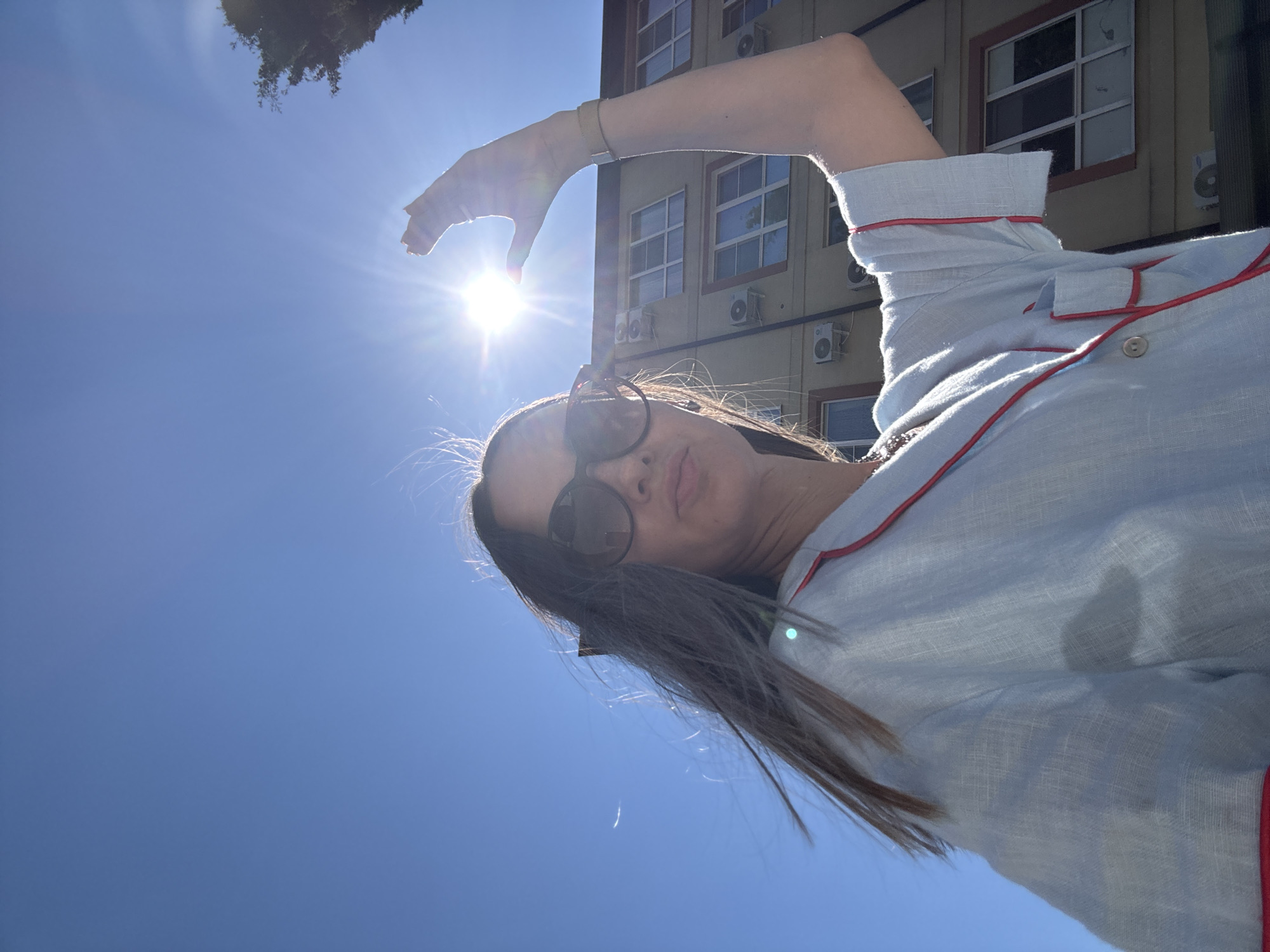 In this sun-drenched selfie, a woman with long, dark hair, wearing oversized sunglasses and a light blue pajama-style top with red trim, poses outdoors. She raises her right hand, her fingers partially obscuring the bright sun, which is positioned high in a clear blue sky. The woman appears to be standing in front of a light-colored building with multiple windows and air conditioning units. In the background, a tree with dark green foliage is visible on the right side of the frame. The overall scene suggests a warm, sunny day in an urban environment, likely Sochi, Russia, given the specific location information. The low angle of the shot emphasizes the bright sky and the sun's intensity.