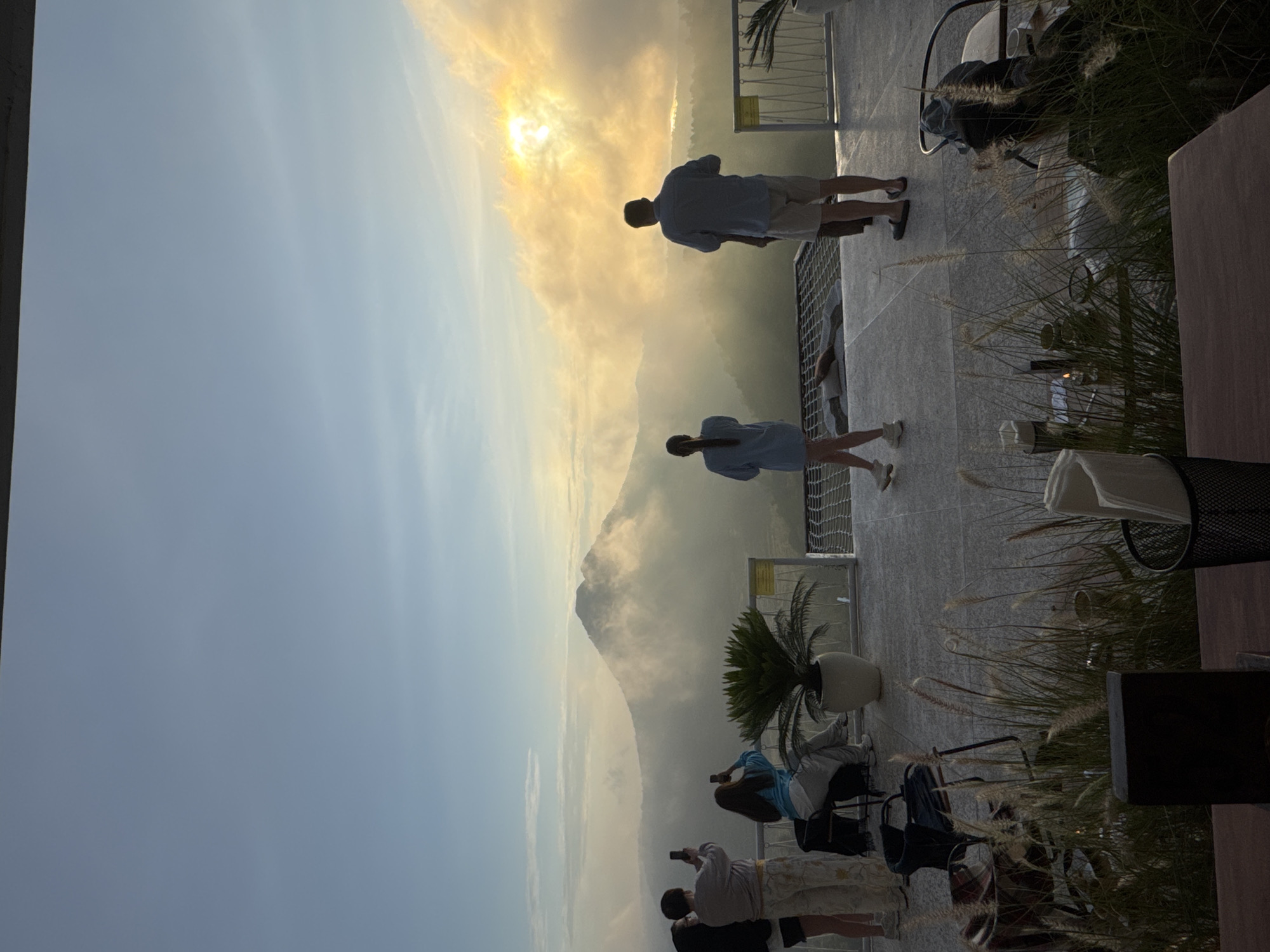 Kedisan, Indonesia. The image depicts a scenic view of a mountainous landscape partially obscured by fog or mist, with a bright sun visible in the sky. In the foreground, there is an elevated terrace or patio with several people. One man and one woman are standing near a railing looking out at the view. A group of people are taking photos. There are plants in pots and chairs and tables. The sky is clear blue above the foggy mountains.
