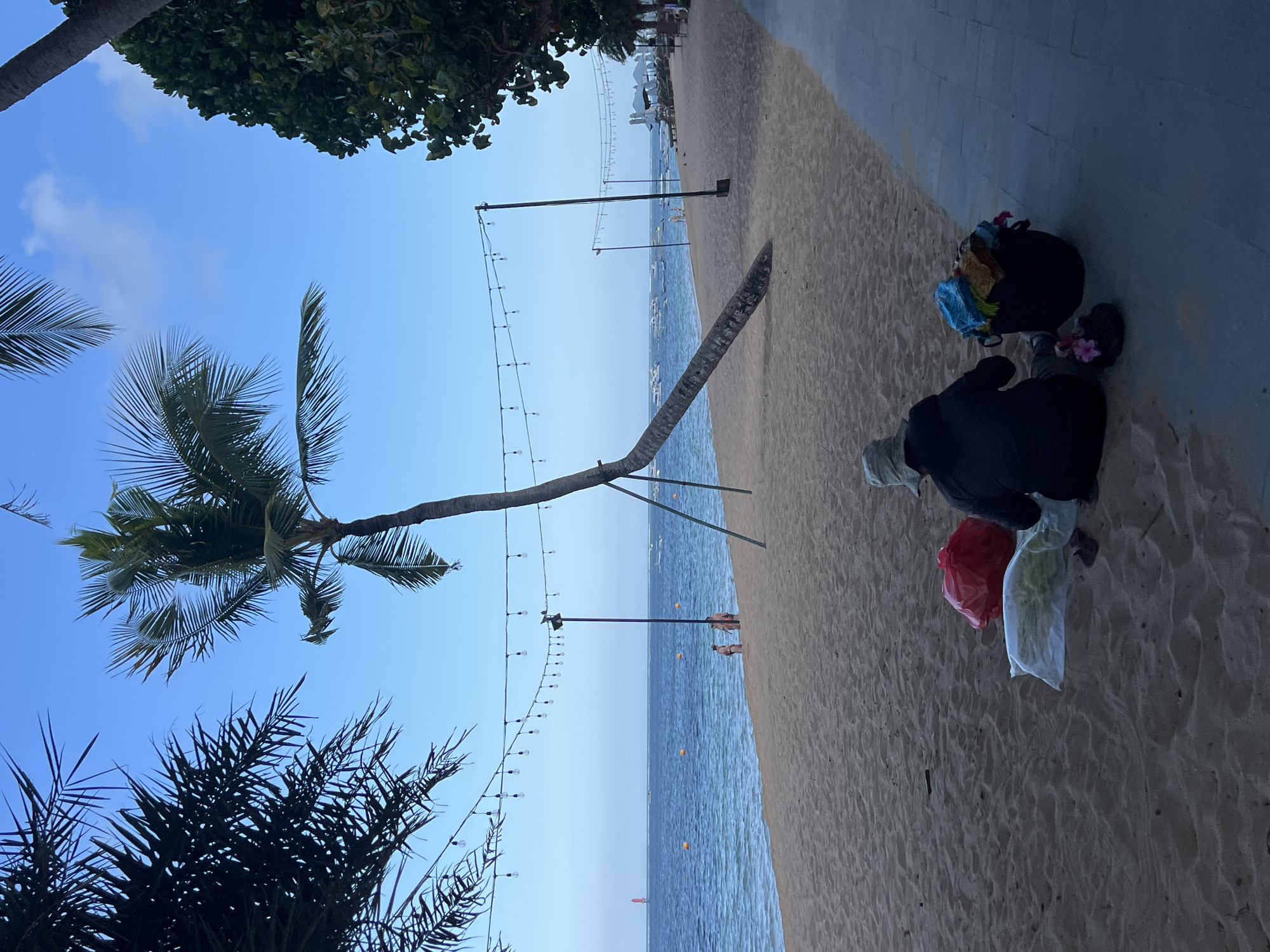 A person sits on a sandy beach in Denpasar, Indonesia, facing a body of water. The person is wearing a hat and a dark jacket, and their belongings, including a red bag and a black basket filled with colorful items, are spread around them. The beach is lined with palm trees and a paved walkway. In the distance, there are several boats on the water, and a few people can be seen wading near the shore. The sky is clear and blue, with a few faint clouds. String lights are visible overhead, spanning across the beach. The overall scene depicts a peaceful day at the beach.