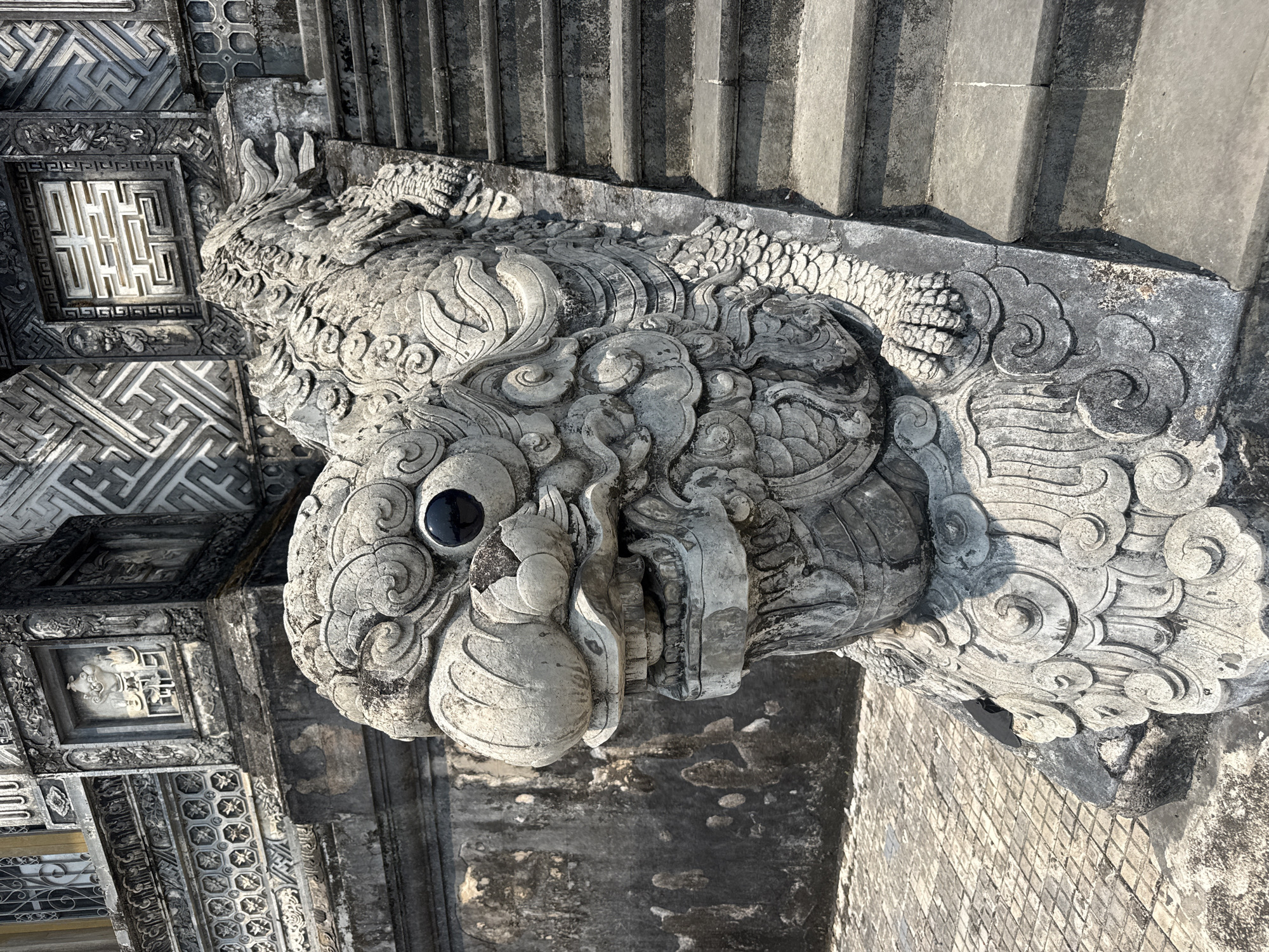 A detailed stone carving of a mythical creature, possibly a dragon-fish hybrid, sits on a stone structure in Thủy Xuân Ward, Vietnam. The carving is gray, weathered, and features intricate details, including scales, swirling patterns, and a prominent eye. The creature's mouth is slightly open, revealing teeth. The carving is positioned on the edge of a stone structure, likely a staircase or platform, which also features decorative patterns and carvings. The surrounding environment consists of stone structures with geometric patterns and textures, suggesting a historic or religious site.