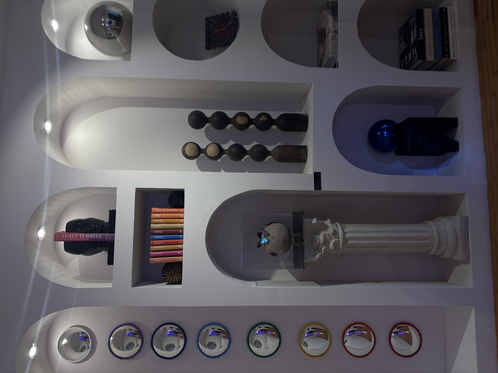 The image shows a built-in shelf in Mexico City, Mexico. The shelf is painted white and has several nooks and crannies in which objects are placed, including balls, sculptures, a book titled "James Turrell A Retrospective", a stack of books related to art, and reflective mirrors in various colors, including blue, green, and yellow. There is also an ancient-looking pillar or column in the center, to the right. The shelf is illuminated by spotlights on the top. The overall impression is of a modern and stylish display of art and decor in a home or gallery.