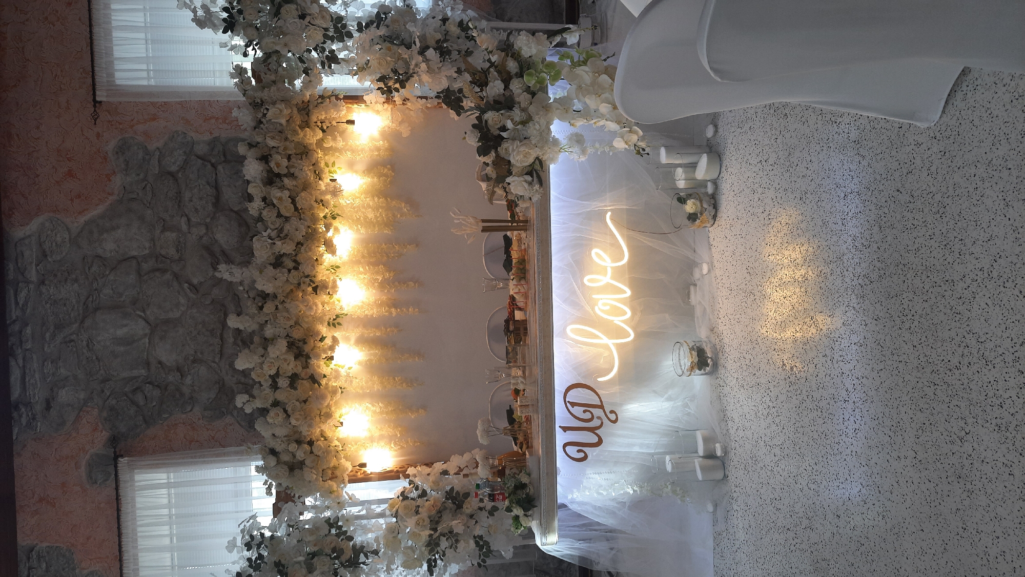 In an indoor event space in Жодзішкаўскі сельскі Савет, Belarus, a long table is elaborately decorated as a sweetheart table or head table for a wedding reception. The table is draped with white tulle fabric, creating a flowing, ethereal effect. Centered on the front of the table is a neon sign that reads "UD love" in a warm, glowing yellow light, with "UD" in a stylized wooden cutout font. The table itself is laden with floral arrangements, glassware, and what appears to be a charcuterie spread.
Above the table, a large, cascading floral arrangement of white roses and other white flowers is suspended, interwoven with delicate white hanging decorations and illuminated by a series of warm pendant lights. The backdrop features a rustic stone wall, partially covered by a textured peachy-pink paint, with two large white-curtained windows visible on either side.
The foreground shows a speckled, light-colored floor where the reflection of the neon sign is visible. Several cylindrical white candles and glass vases containing white flowers and decorative elements are placed on the floor in front of the table, adding to the romantic ambiance. Two chairs draped in white fabric are positioned to the right of the frame, suggesting they are for guests or the couple seated at the table. The overall impression is one of elegant and romantic wedding decor.