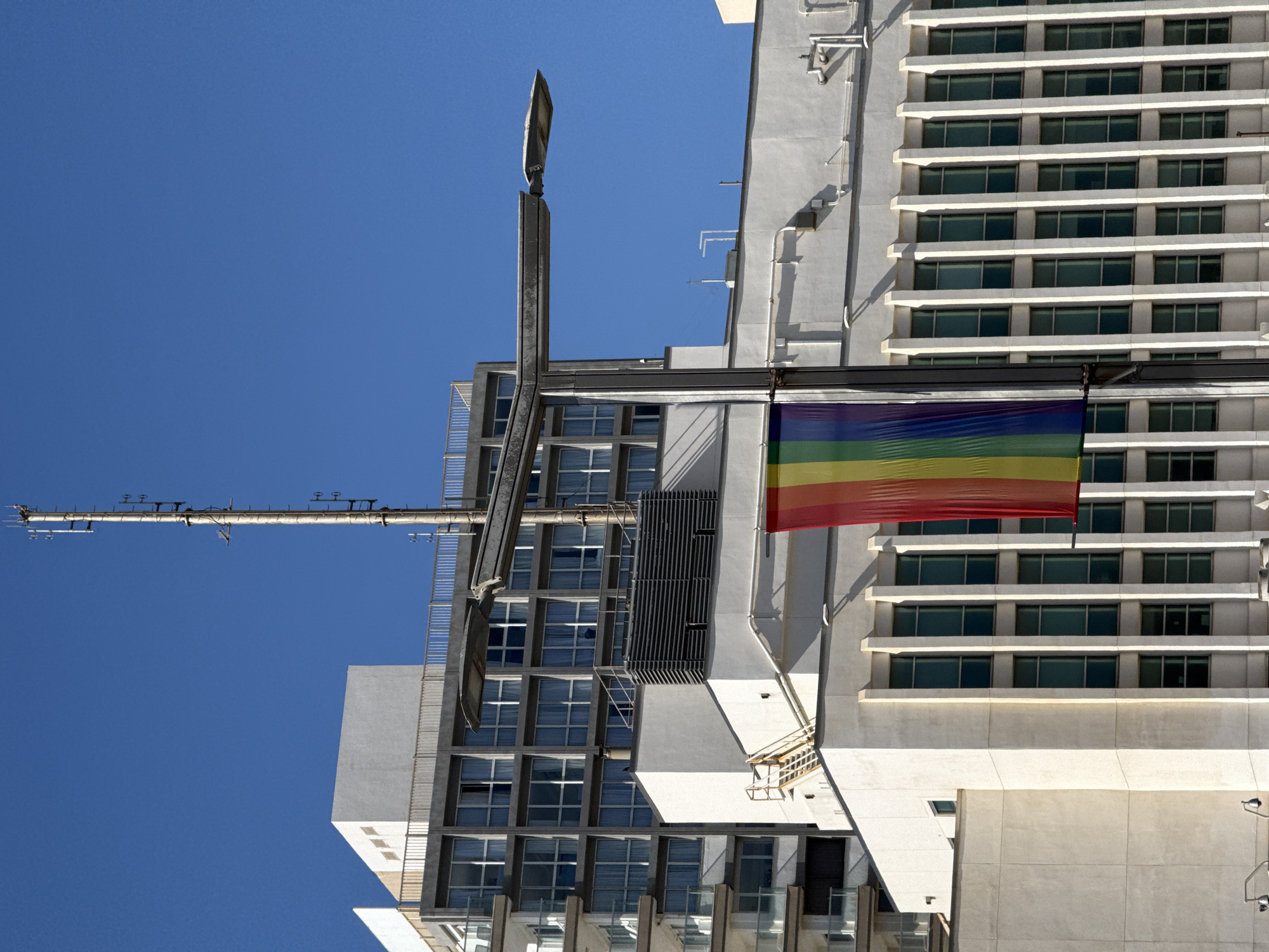 A low-angle shot of buildings in Tel Aviv, Israel under a clear blue sky, featuring a prominent rainbow flag suspended between structures. The architecture is modern, with a mix of white concrete and glass windows. Visible details include a lamppost, pipes running along the building facades, and what appears to be an antenna extending from one building. The rainbow flag is brightly colored and prominently displayed.