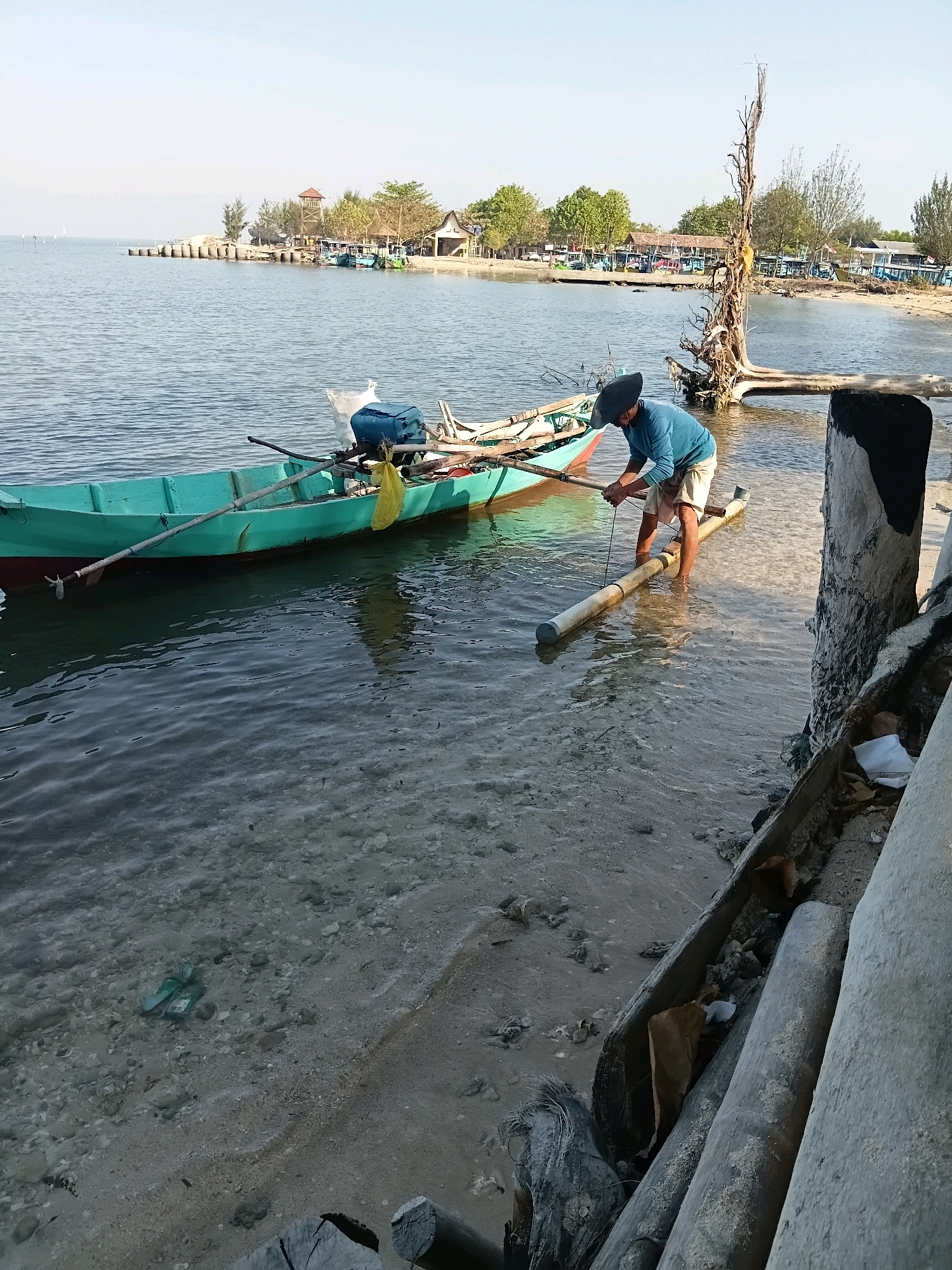 A man wearing a blue long-sleeved shirt and light-colored shorts, and a wide-brimmed hat, stands in shallow water next to a turquoise boat. He is holding a pole in each hand, working with a white pipe-like object that is partially submerged in the water. The boat is moored and contains oars, a yellow bag, and a blue container. The water is clear, with sand and pebbles visible on the bottom. In the background, a sandy beach is lined with trees and buildings. Further in the distance, several other boats are visible in the water, and a watchtower stands on a small island. The scene appears to be a waterfront area in Cikal, Indonesia, possibly a fishing village or harbor. The lighting suggests it is either early morning or late afternoon.