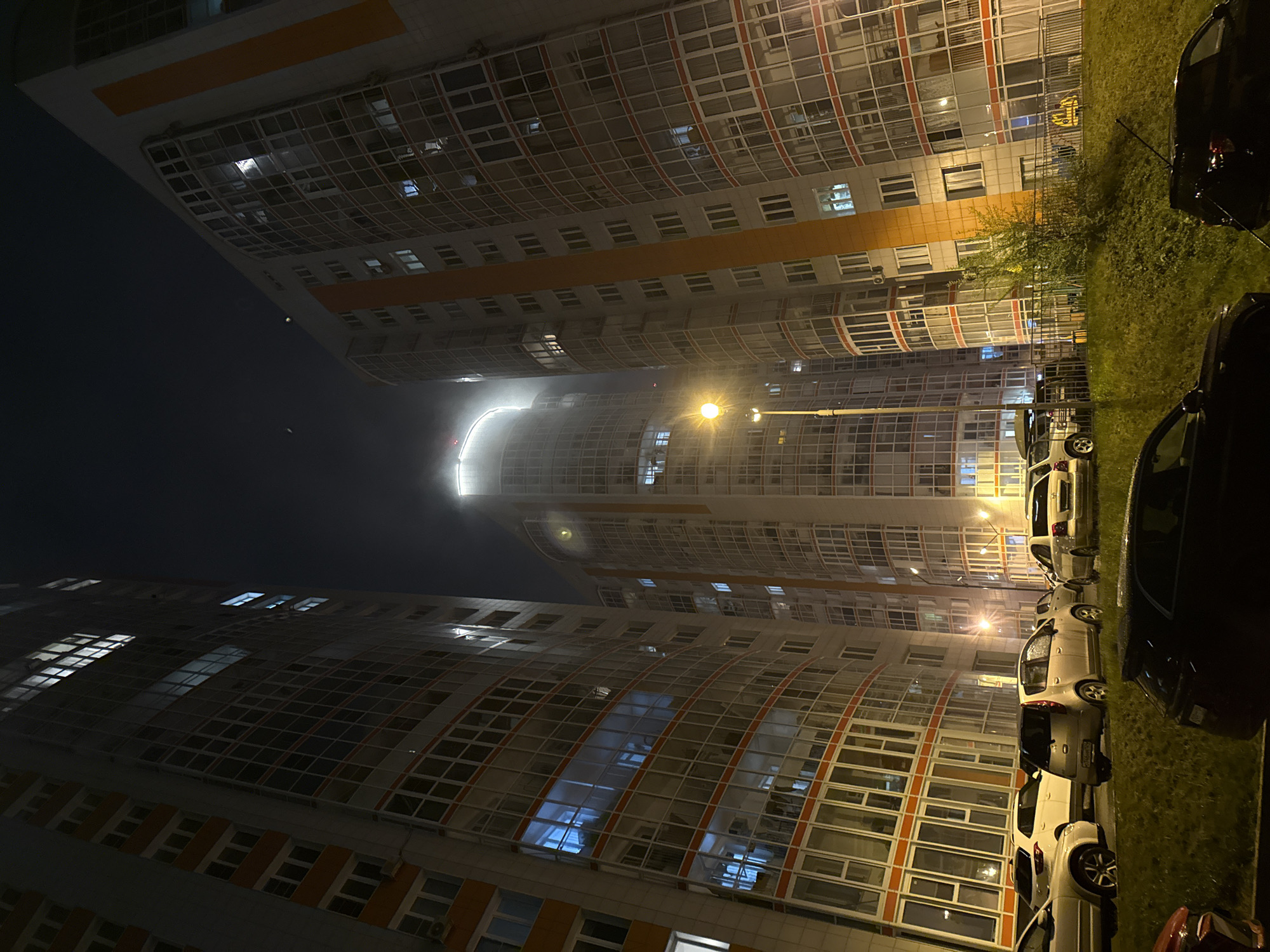 The image depicts a night scene in Krasnoyarsk, Russia, featuring two tall apartment buildings with curved facades facing each other. Windows are visible across both structures, with some illuminated, suggesting occupancy. A street lamp provides artificial light, illuminating the space between the buildings. Cars are parked along the side of one building, situated on a grass area. The sky is dark and partially obscured by fog.