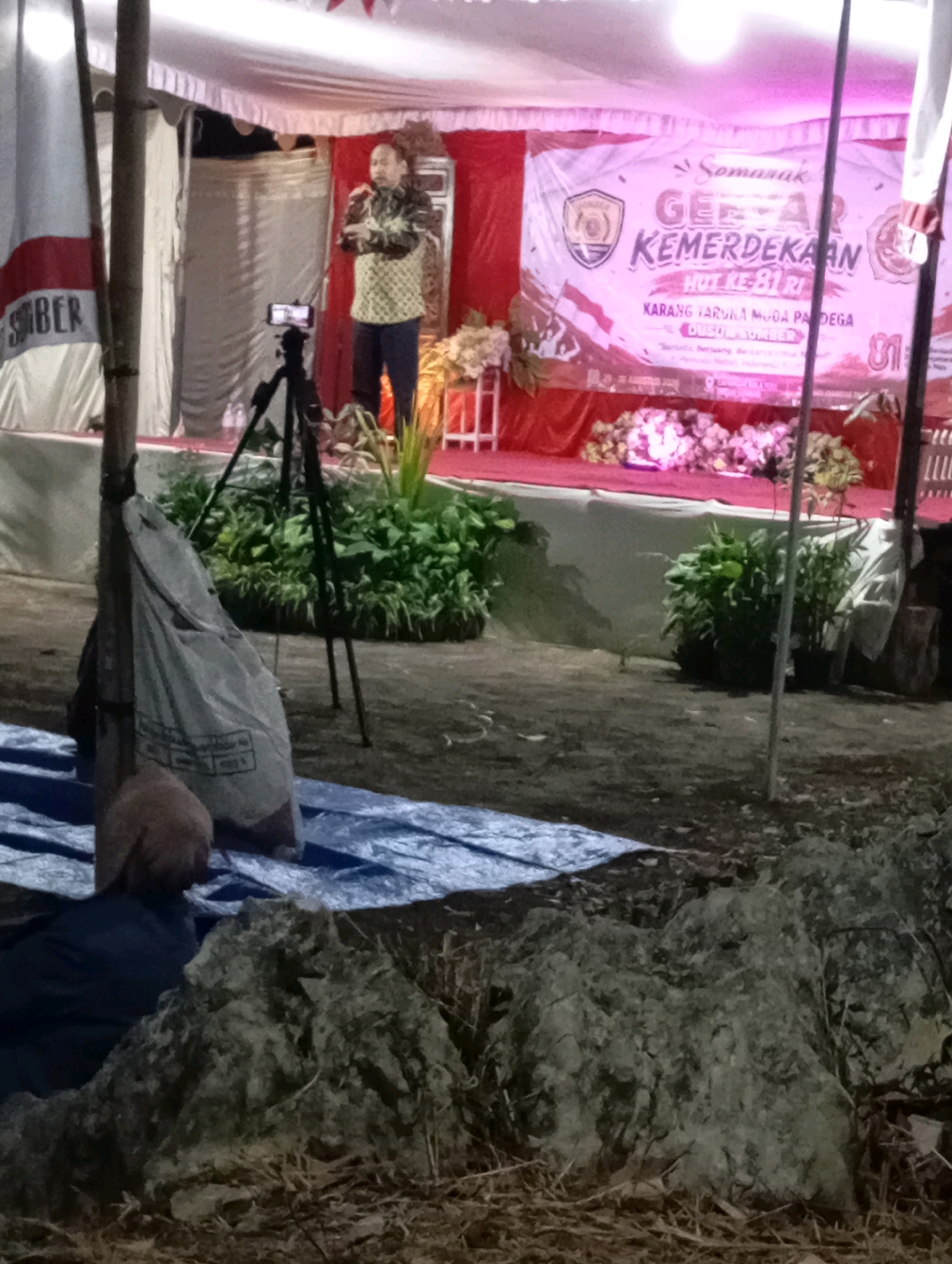 The image depicts an outdoor event, likely a ceremony or presentation, taking place in the city of Wonogondo, Indonesia. A man, wearing a patterned shirt and dark pants, is standing on a raised stage, speaking into a microphone. He appears to be addressing an audience, although the audience is not visible. In front of the stage, a tripod with a smartphone mounted on it is set up, suggesting that the event is being recorded or live-streamed. A large banner with Indonesian text and graphics is displayed behind the speaker, indicating the theme of the event is "Gelar Kemerdekaan HUT KE-81" (Grand Celebration of Independence 81st Anniversary). The stage is decorated with red curtains and floral arrangements. To the left of the stage, a flag with text is visible. In the foreground, a blue tarp covers the ground, and a large grey bag is placed on it, tied to a wooden pole. Rocks and dried vegetation are scattered on the ground in the lower portion of the image. The lighting suggests it is evening or night, with artificial lights illuminating the stage area.