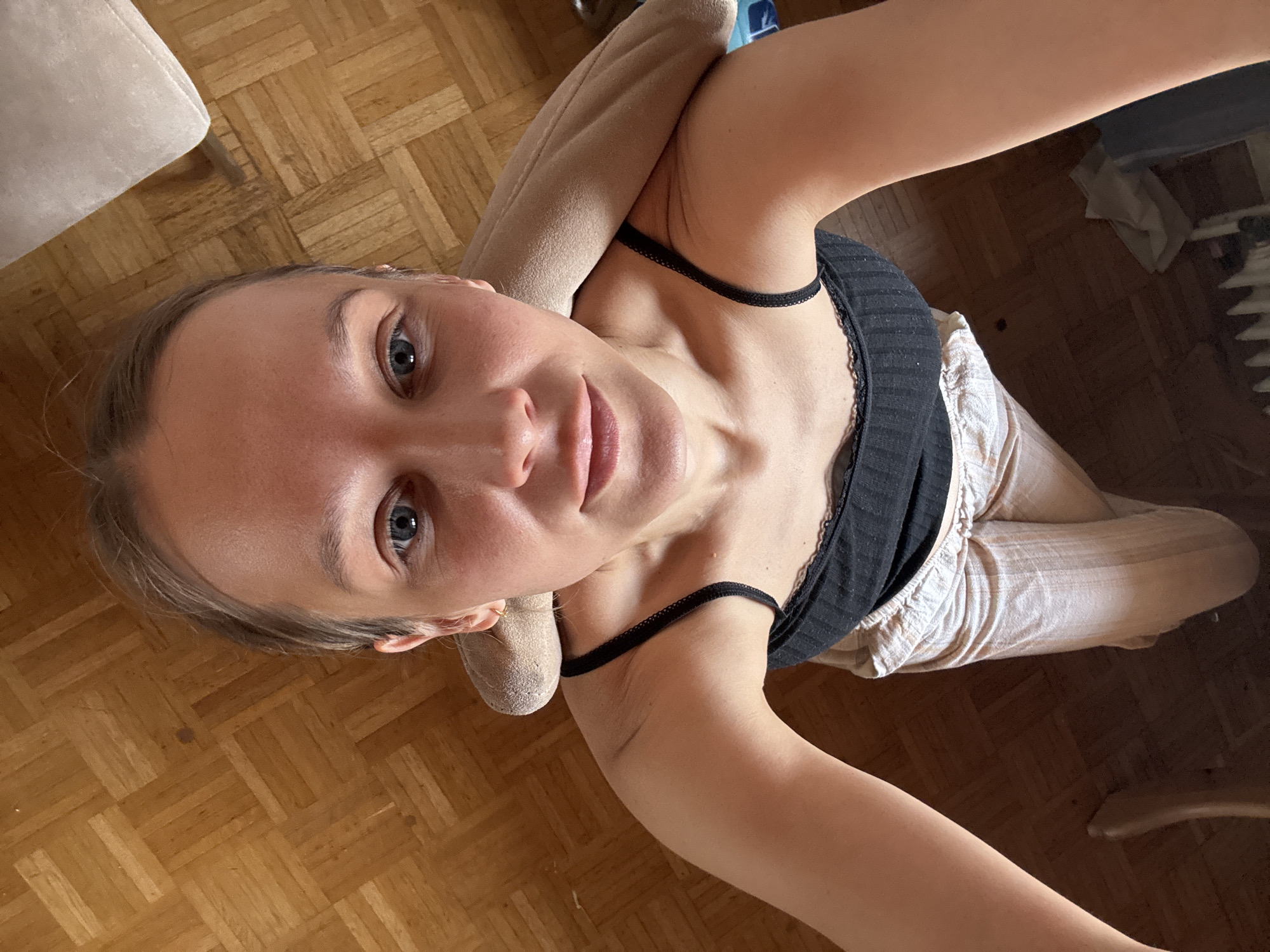 The media file is a selfie of a young woman taken from a high angle. She is lying down, with her head propped up on a light-colored cushion. She is wearing a black tank top with lace trim and light-colored, striped pants. Her hair is blonde and appears to be pulled back. Her eyes are blue and she is looking at the camera with a slight smile.
The scene is indoors, with a wooden parquet floor visible. A portion of a light-colored chair or couch is also visible in the upper left corner of the image. In the background, there is a radiator and some crumpled papers or fabric. The lighting suggests it is daytime, possibly late morning or afternoon. There is no visible text in the image. The woman appears relaxed and comfortable. The location context is Überherrn, Germany.